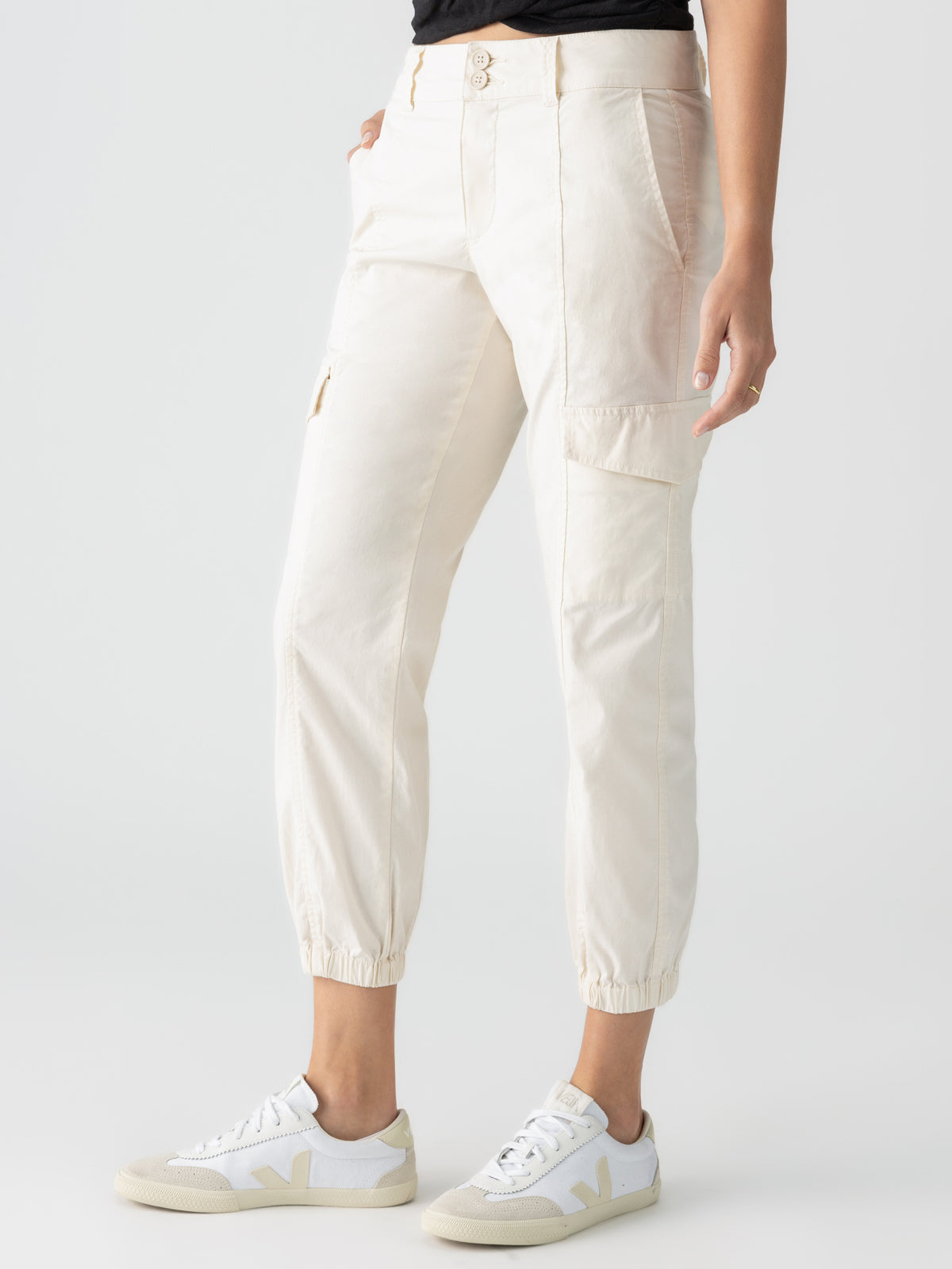 A person wearing the Rebel Pant Eco Natural by Sanctuary Clothing, featuring multiple pockets and cinched cuffs, paired with white sneakers with beige accents. The person is standing against a plain light background with their hands at their sides. The upper body is mostly out of the frame.