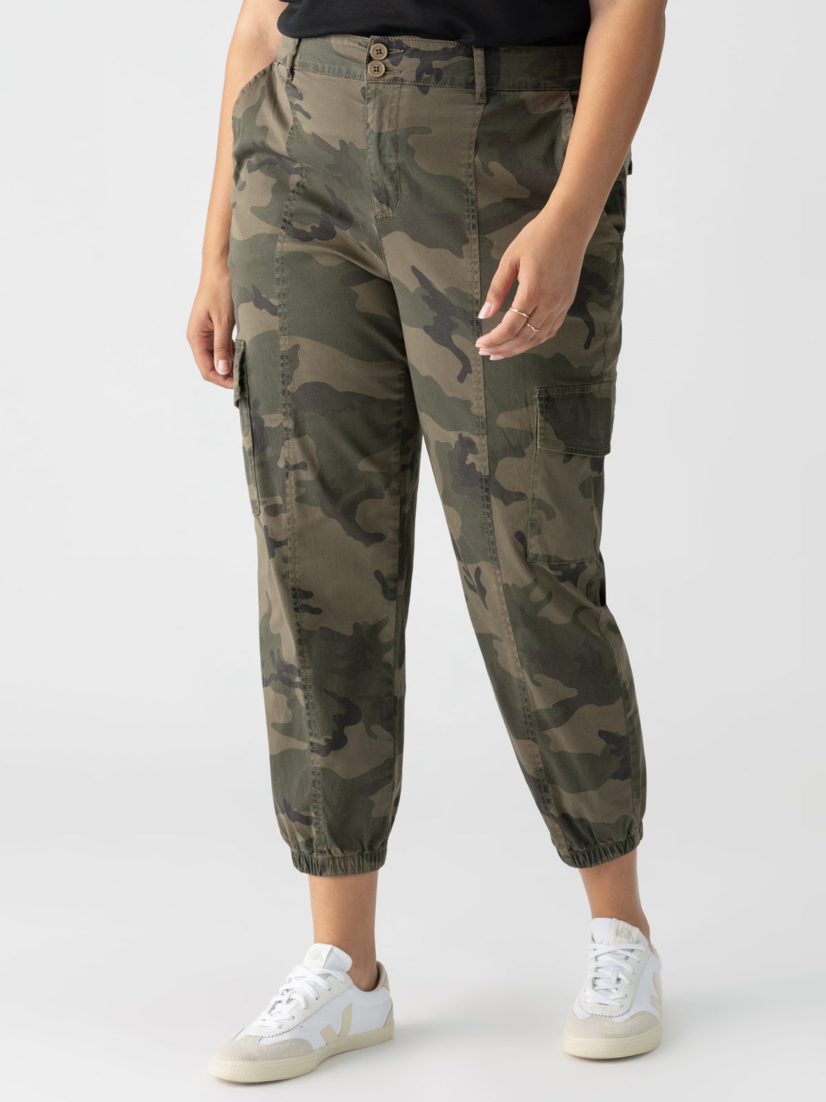 A person is wearing the Rebel Pant Hiker Camo from Sanctuary Clothing's Inclusive Collection, featuring elasticated cuffs, paired with a black top. They are also sporting white sneakers with a light-colored "V" logo on the side. The person is standing against a plain white background.