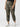 A person is wearing the Rebel Pant Hiker Camo from Sanctuary Clothing's Inclusive Collection, featuring elasticated cuffs, paired with a black top. They are also sporting white sneakers with a light-colored "V" logo on the side. The person is standing against a plain white background.