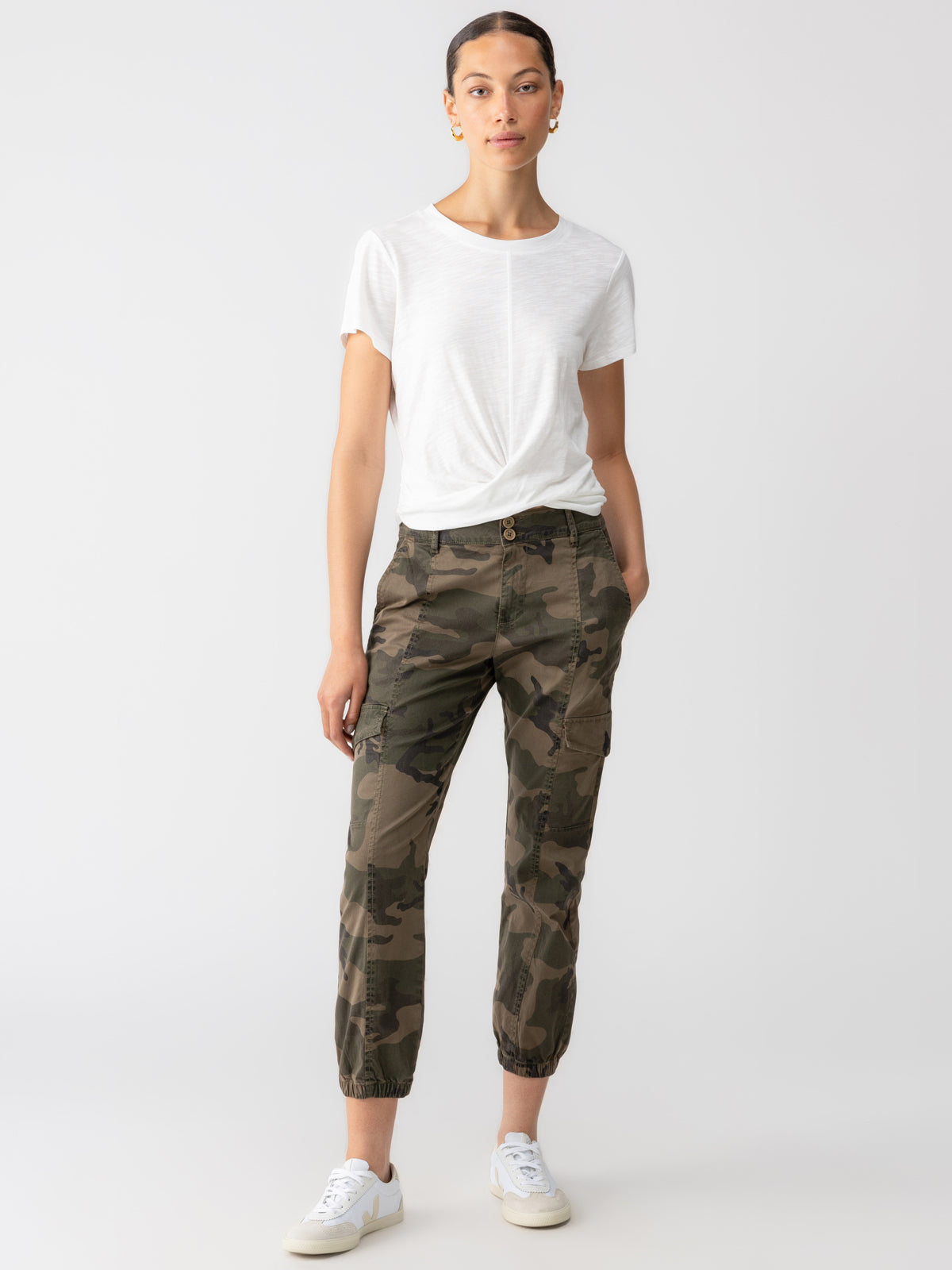 A person stands against a plain background, wearing a white short-sleeve T-shirt and the Rebel Pant Hiker Camo by Sanctuary Clothing. They have white sneakers, hands in their pockets, and hair pulled back. The overall look is casual and minimalist.