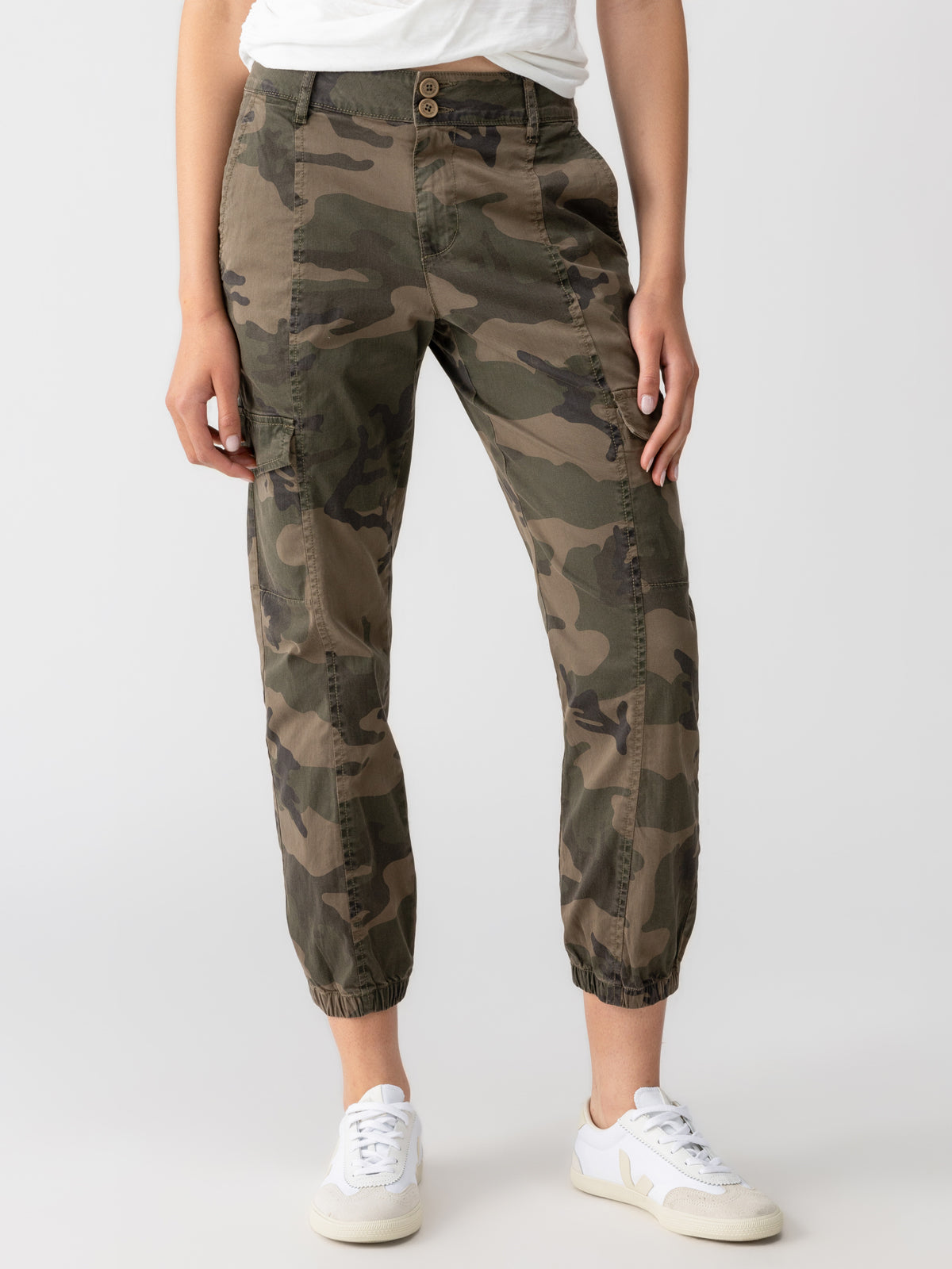 A person wearing the Rebel Pant Hiker Camo from Sanctuary Clothing, paired with a white top and white sneakers. The camouflage pants feature side pockets and elastic cuffs at the ankles. The person is standing against a plain background.