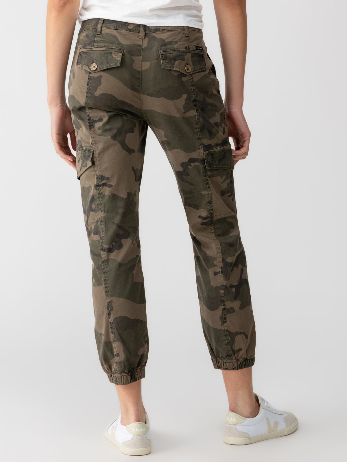 Rear view of a person wearing the Rebel Pant Hiker Camo from Sanctuary Clothing, featuring side pockets and elastic cuffs, paired with white sneakers. The person is dressed in a white top, and the background is plain and light-colored.