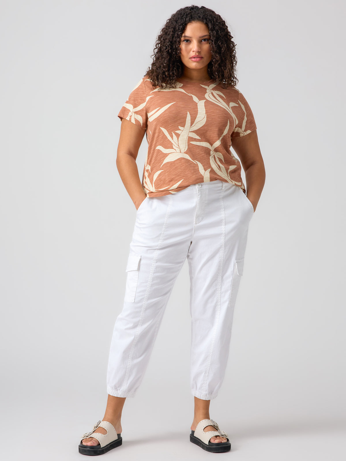 A person with curly hair stands against a plain background, dressed in a short-sleeve, brown top adorned with a leaf pattern. The top is tucked into the Rebel Pant Brilliant White from Sanctuary Clothing's Inclusive Collection. Their hands are casually placed in their pockets, and they complete the look with white sandals.