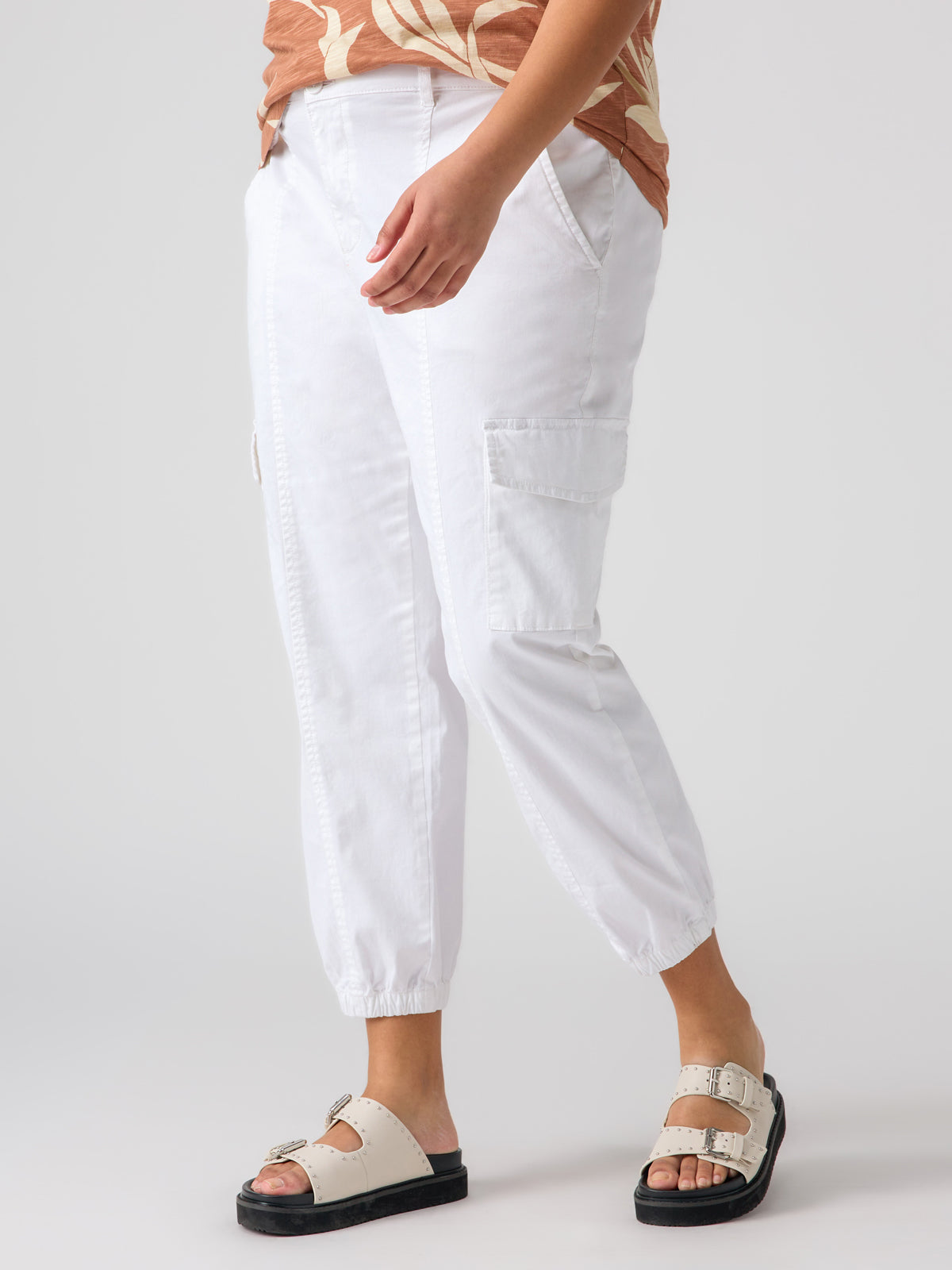 A person wearing a brown-and-beige top is shown from the waist down, dressed in Sanctuary Clothing's Rebel Pant Brilliant White from the Inclusive Collection, featuring side pockets and rolled cuffs, and white sandals with two buckles and black soles. The background is plain white.