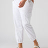 A person wearing a brown-and-beige top is shown from the waist down, dressed in Sanctuary Clothing's Rebel Pant Brilliant White from the Inclusive Collection, featuring side pockets and rolled cuffs, and white sandals with two buckles and black soles. The background is plain white.