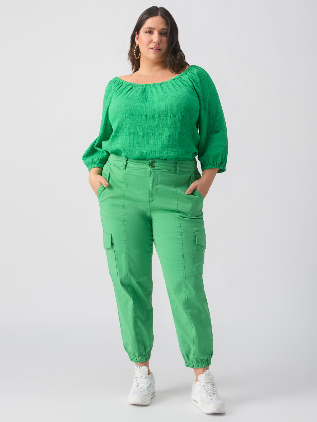 A woman stands confidently against a plain background, wearing a green long-sleeve blouse and the Rebel Pant from the Green Goddess Inclusive Collection by Sanctuary Clothing. She has her hands in her pockets and is looking towards the camera. She is also wearing white sneakers and gold hoop earrings.