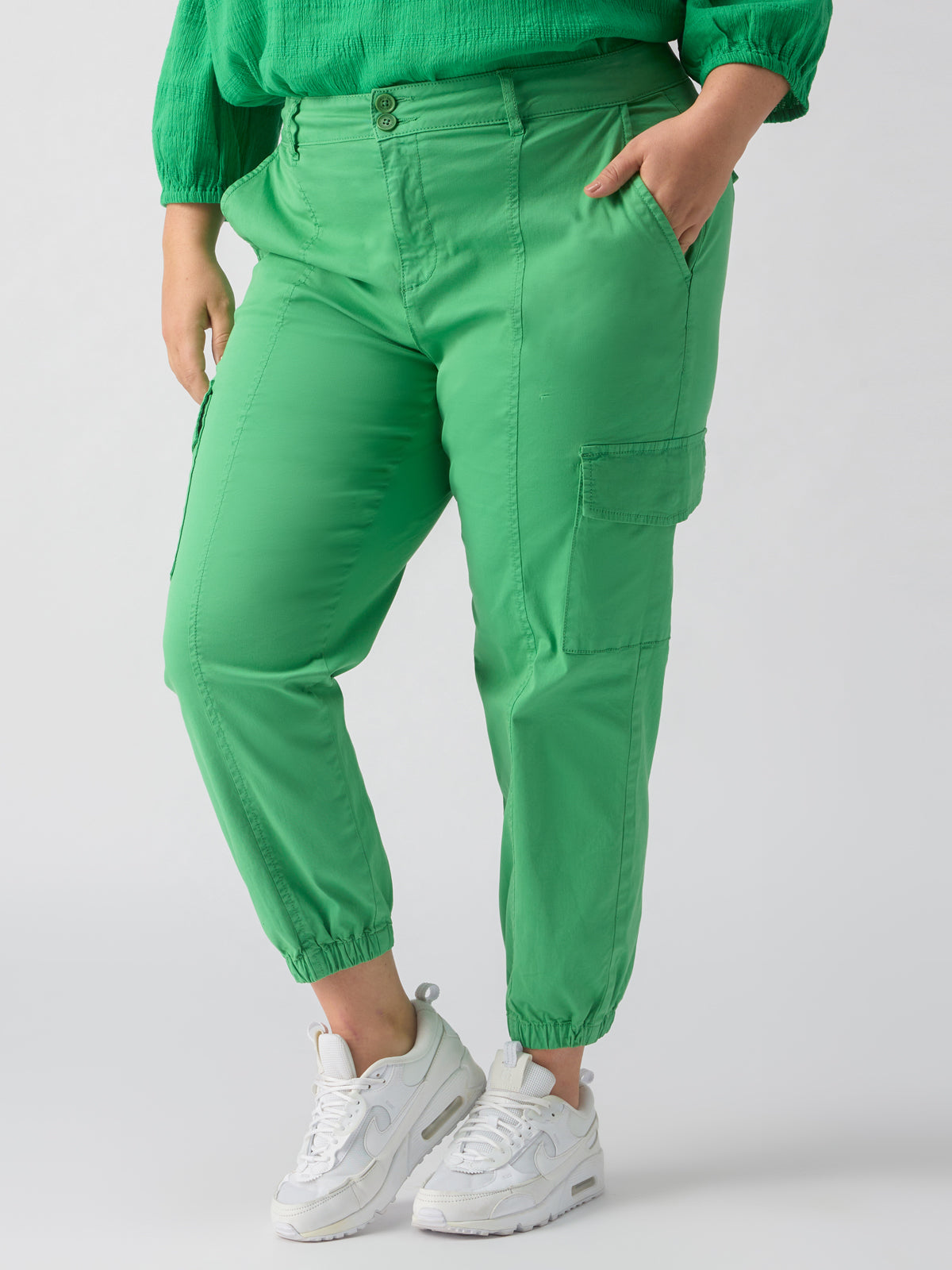 A person is wearing the Rebel Pant Green Goddess from Sanctuary Clothing's Inclusive Collection, paired with white sneakers. One hand is in the pocket of the bright green cargo pants with an elasticated cuff at the ankles, and they are also wearing a long-sleeved green top. The background is plain and light-colored.