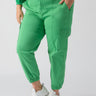 A person is wearing the Rebel Pant Green Goddess from Sanctuary Clothing's Inclusive Collection, paired with white sneakers. One hand is in the pocket of the bright green cargo pants with an elasticated cuff at the ankles, and they are also wearing a long-sleeved green top. The background is plain and light-colored.