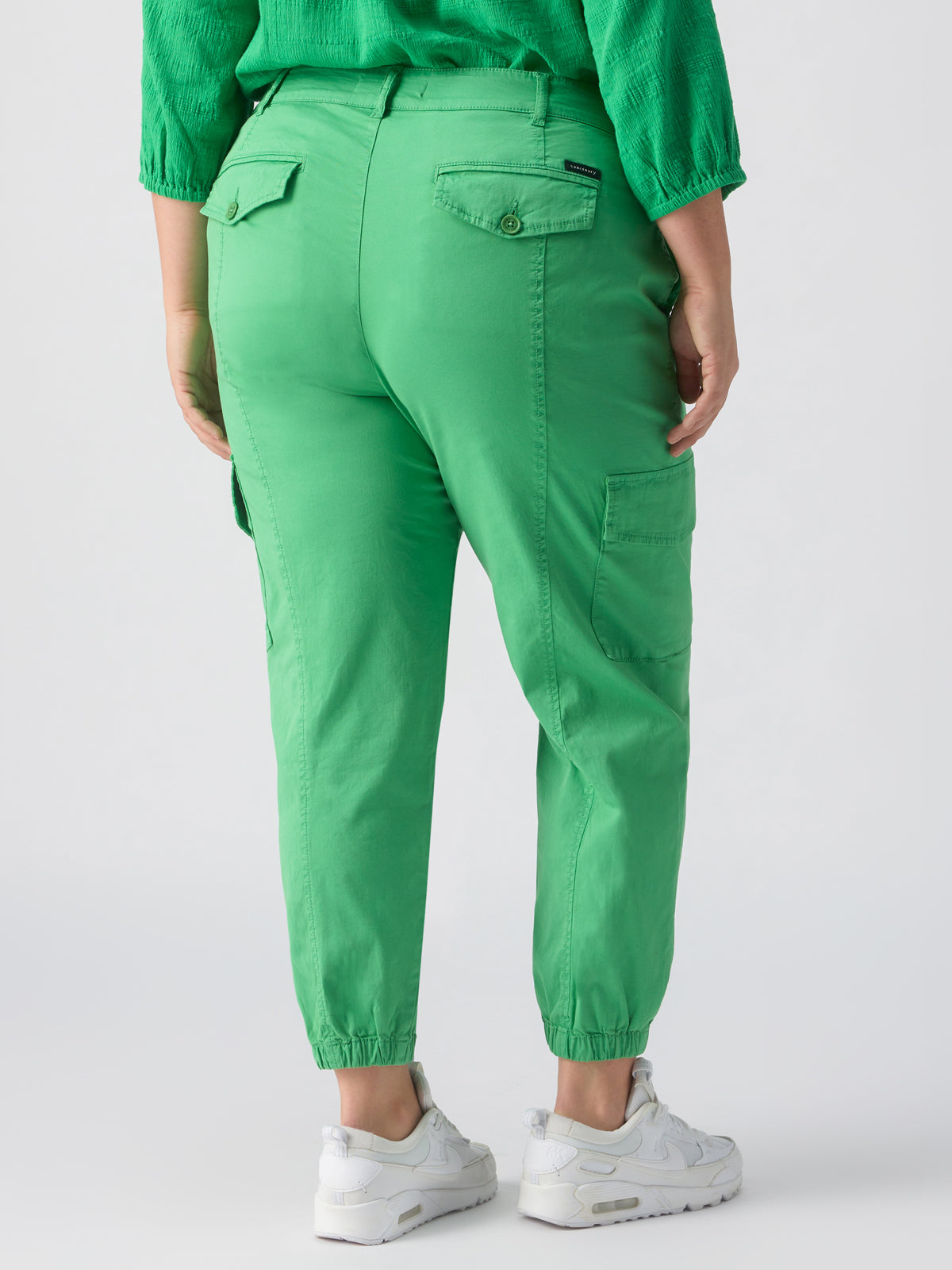 A person is wearing the Rebel Pant in Green Goddess from the Inclusive Collection by Sanctuary Clothing. The bright green cargo pants feature multiple pockets and elastic cuffs at the ankles. They are also wearing a green long-sleeve top and white sneakers. The image shows the person from the back, highlighting the fit and style of these distinctive pants.