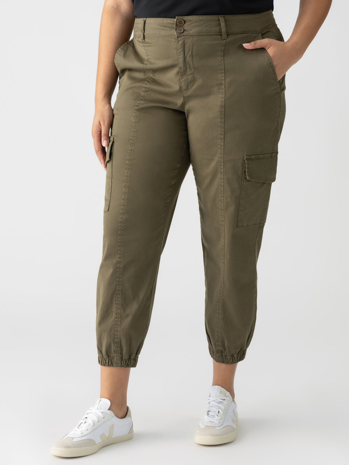 A person is wearing the Rebel Pant Hiker Green from Sanctuary Clothing's Inclusive Collection with a black top. The pants feature multiple pockets, including a side pocket on the thigh, and are cinched at the ankles. The person is also wearing white sneakers against a plain white background.