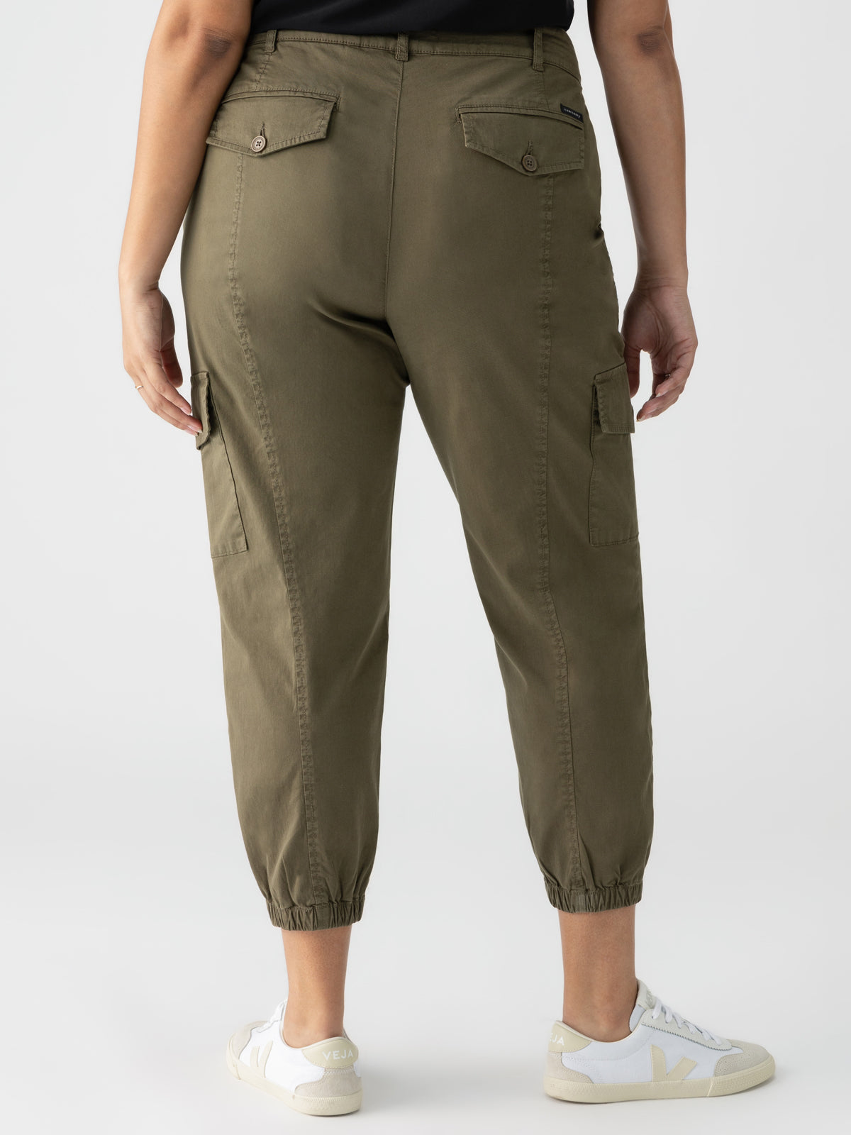 A person is standing with their back to the camera, wearing the Rebel Pant Hiker Green from Sanctuary Clothing's Inclusive Collection, featuring elasticated cuffs and two back pockets with buttoned flaps. They are also sporting white sneakers with light-colored details and a black top, all set against a plain white background.