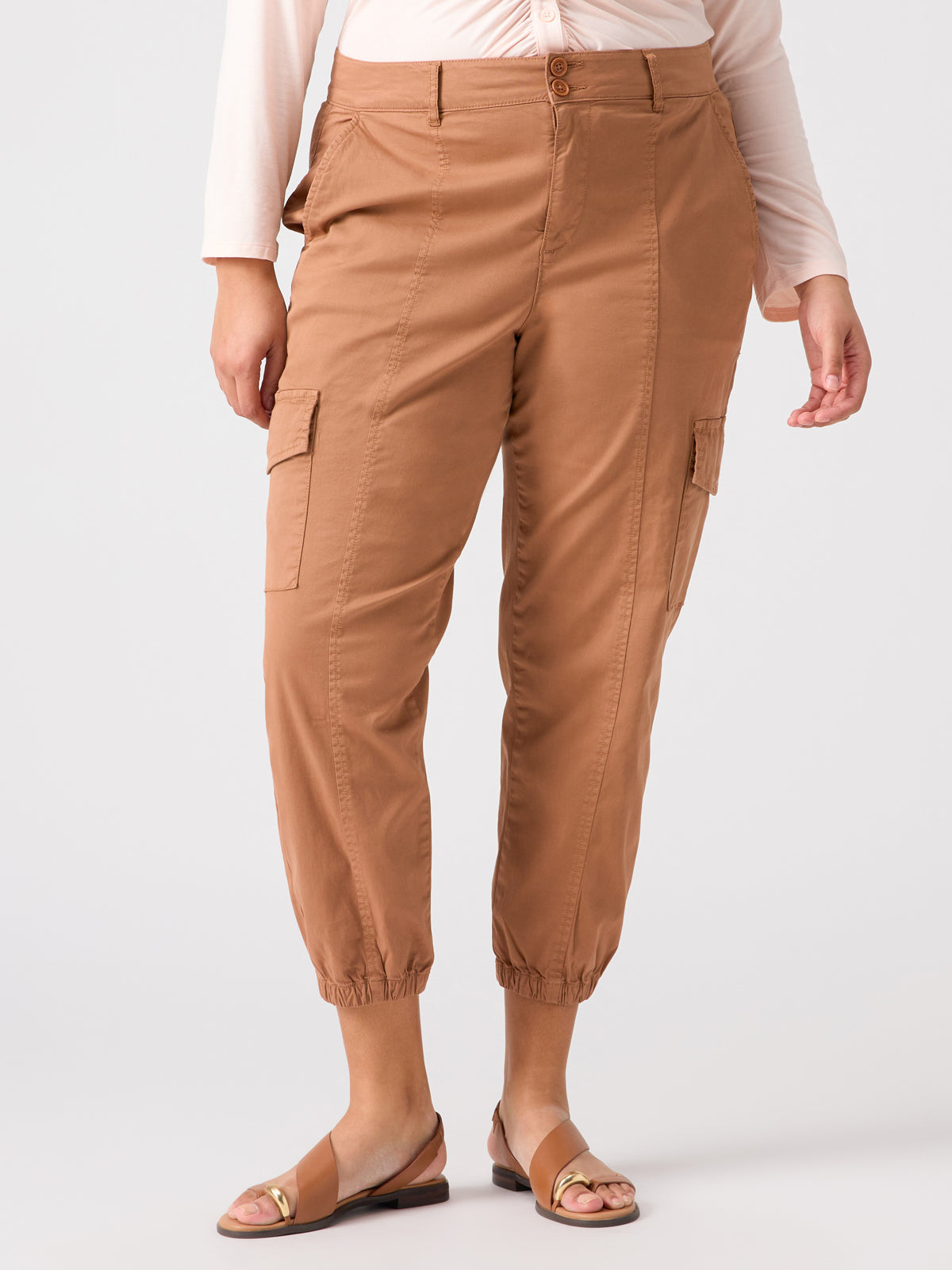 A person wearing the Rebel Pant Mocha Mousse from Sanctuary Clothing's Inclusive Collection with elastic cuffs, a light long-sleeve top, and brown sandals. The pants feature multiple pockets and button closures at the waistband. The background is plain and light-colored.