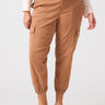 A person wearing the Rebel Pant Mocha Mousse from Sanctuary Clothing's Inclusive Collection with elastic cuffs, a light long-sleeve top, and brown sandals. The pants feature multiple pockets and button closures at the waistband. The background is plain and light-colored.