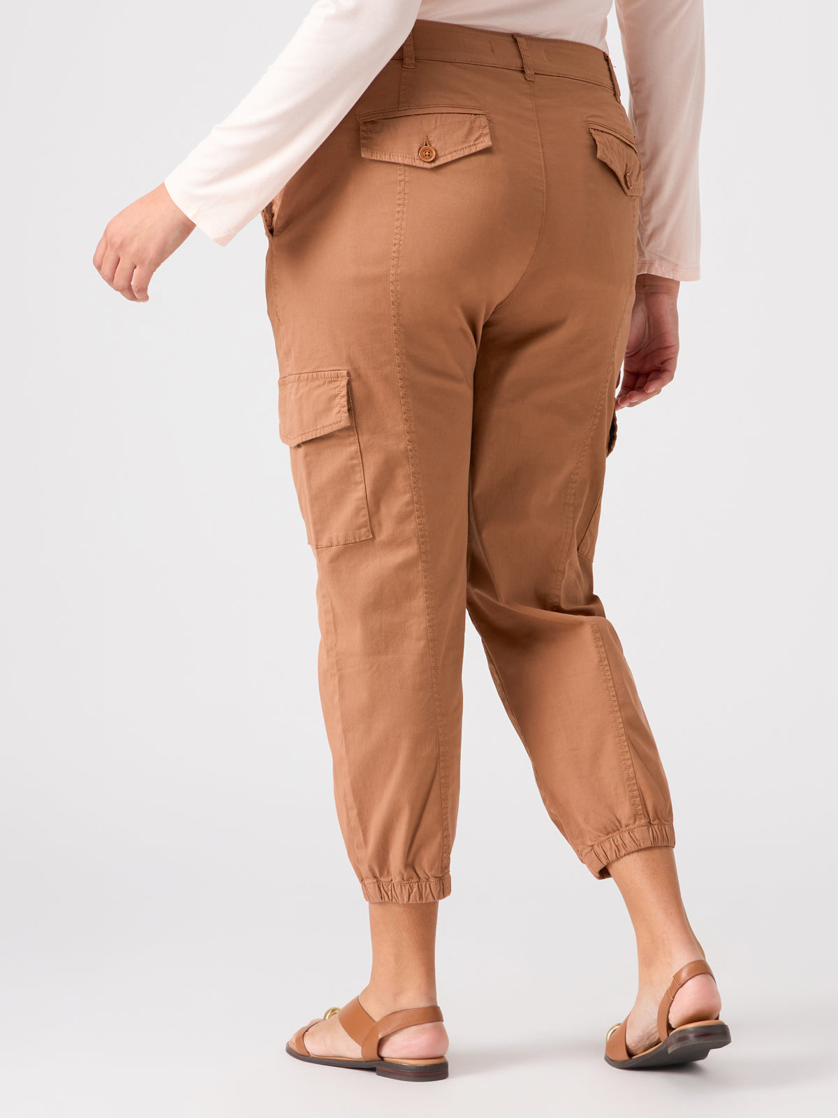 A person is wearing the Rebel Pant Mocha Mousse from the Inclusive Collection by Sanctuary Clothing. The pants feature elastic cuffs and pockets on the sides and back. They are also wearing a long-sleeved white shirt and brown sandals. The background is plain white, and the person is standing with one foot forward, facing away.