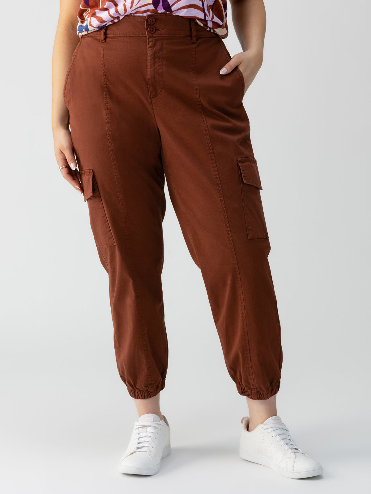 A person stands wearing Sanctuary Clothing's Rebel Pant Rich Clay from the Inclusive Collection and a patterned top. Their left hand is in a pocket, while their right arm rests down by their side. They are wearing white sneakers. The background is plain and unfocused.