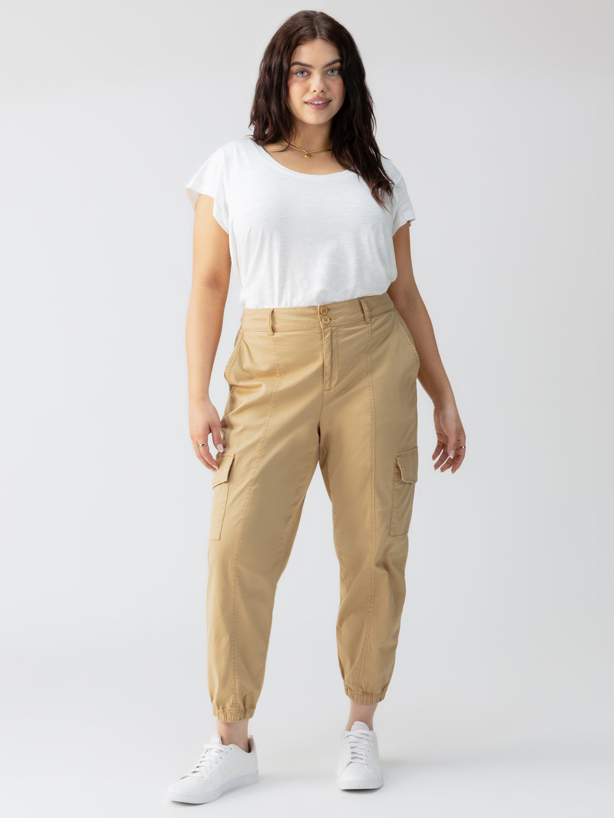 A woman with long dark hair stands confidently against a plain background, wearing a white t-shirt, the Rebel Pant True Khaki from Sanctuary Clothing's Inclusive Collection with pockets, and white sneakers. She smiles slightly and has her hands relaxed by her sides.