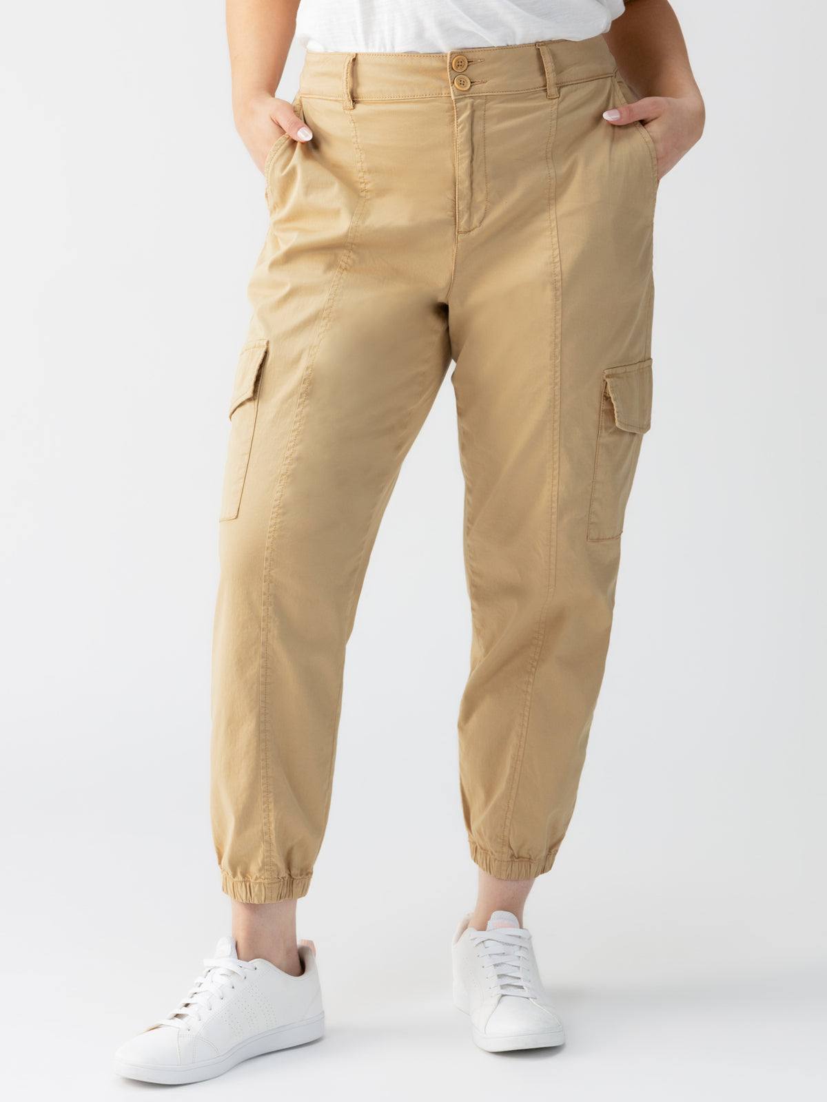 Person wearing the Rebel Pant True Khaki from Sanctuary Clothing's Inclusive Collection, featuring elastic ankle cuffs, side pockets, and button details at the waist. They are paired with a white t-shirt and white sneakers, with their hands tucked into the pants' pockets against a plain white background.