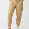 Person wearing the Rebel Pant True Khaki from Sanctuary Clothing's Inclusive Collection, featuring elastic ankle cuffs, side pockets, and button details at the waist. They are paired with a white t-shirt and white sneakers, with their hands tucked into the pants' pockets against a plain white background.