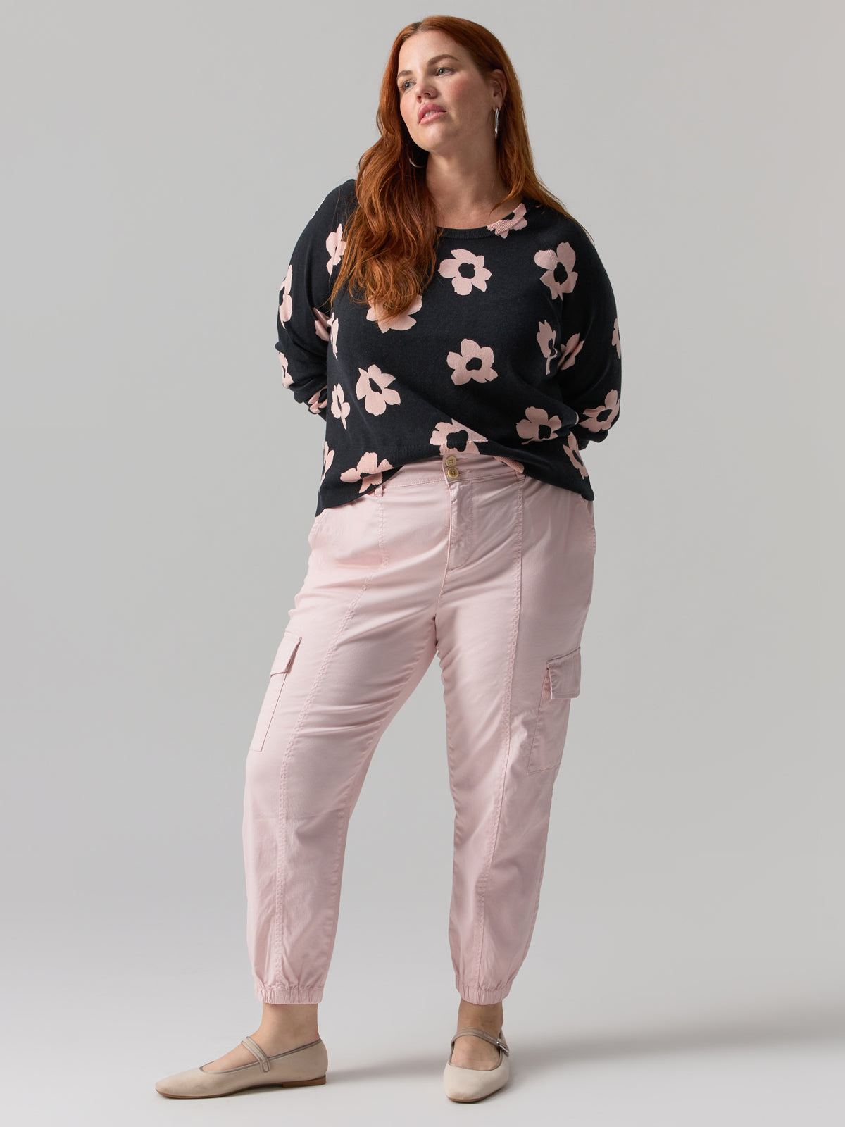 A woman with long red hair poses confidently against a gray background. She is wearing a black sweater adorned with pink floral patterns, the Sanctuary Clothing Rebel Pant in Washed Pink No 3 from their Inclusive Collection, and beige flats. Her hands rest casually in her pockets, and she has a slight, relaxed smile.