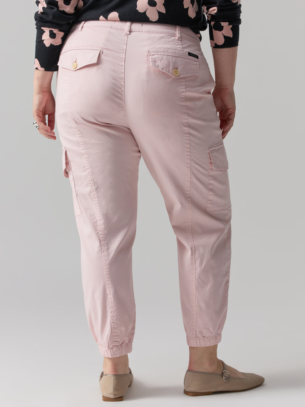 A person is standing with their back to the camera, clad in Sanctuary Clothing's Rebel Pant Washed Pink No 3 from the Inclusive Collection, featuring elasticized ankles and buttoned back pockets. They are also wearing a black long-sleeve shirt adorned with a pink flower pattern and beige flat shoes.