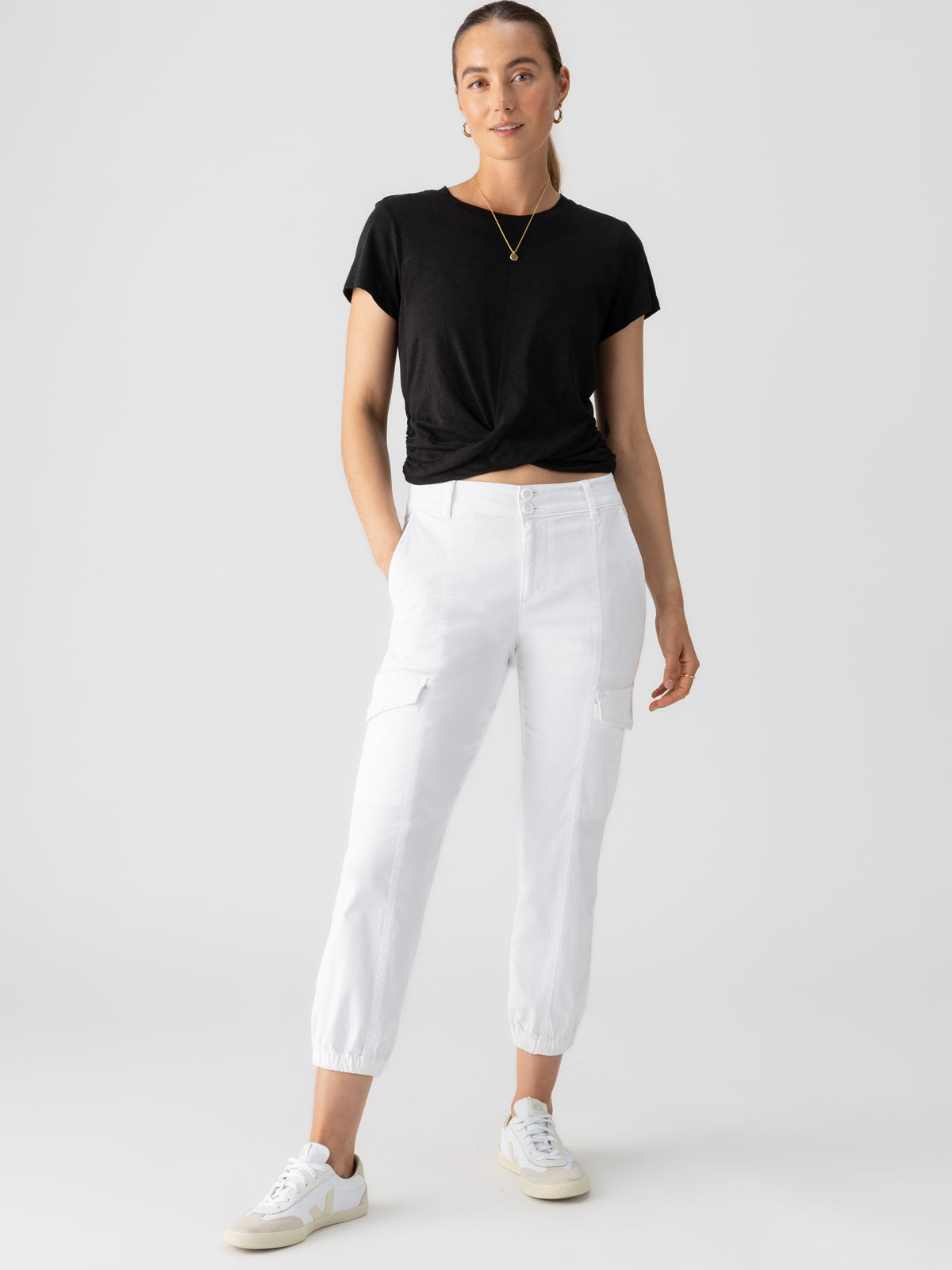 A person stands confidently against a plain background, wearing a black t-shirt tucked into the Rebel Pant Brilliant White by Sanctuary Clothing and white sneakers. The person has their hair tied back and accessorizes with a gold necklace. The overall look is casual and stylish.