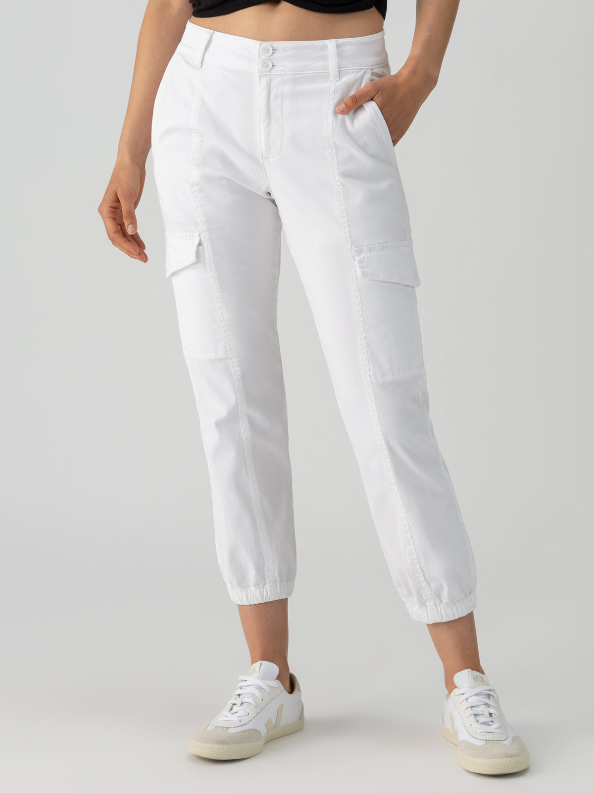 A person wearing the Rebel Pant in Brilliant White by Sanctuary Clothing, featuring a high waist, button and zip closure, and multiple pockets including side flap pockets. The pants are paired with white and beige sneakers. The person has one hand in a pocket and is standing against a light gray background.