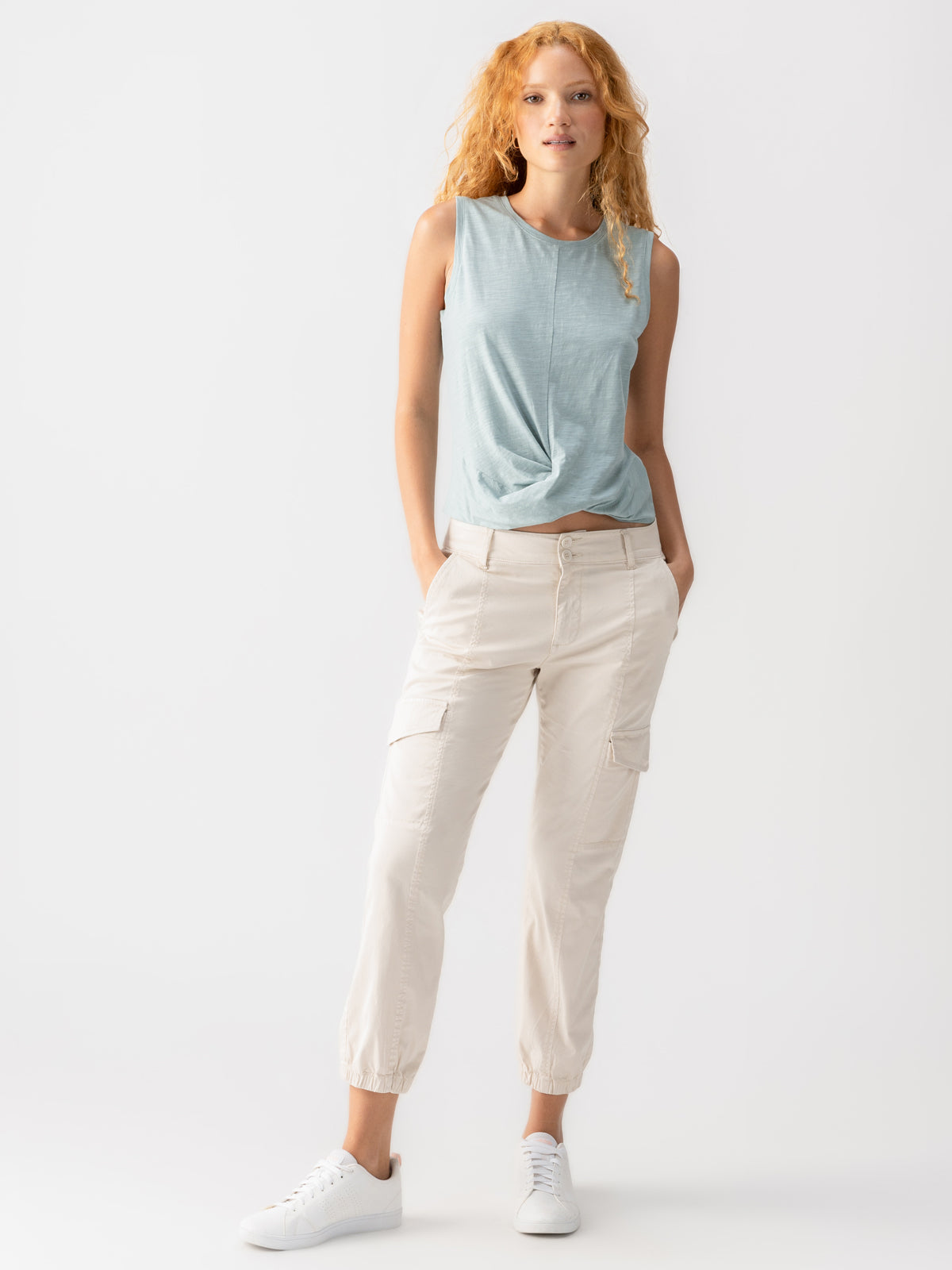 A person with long, wavy red hair wears a light blue sleeveless top with a front knot and the Rebel Pant Crystal Stone by Sanctuary Clothing, which are cream-colored cargo pants. They are also wearing white sneakers. They stand against a plain white background with hands in pockets, looking forward.