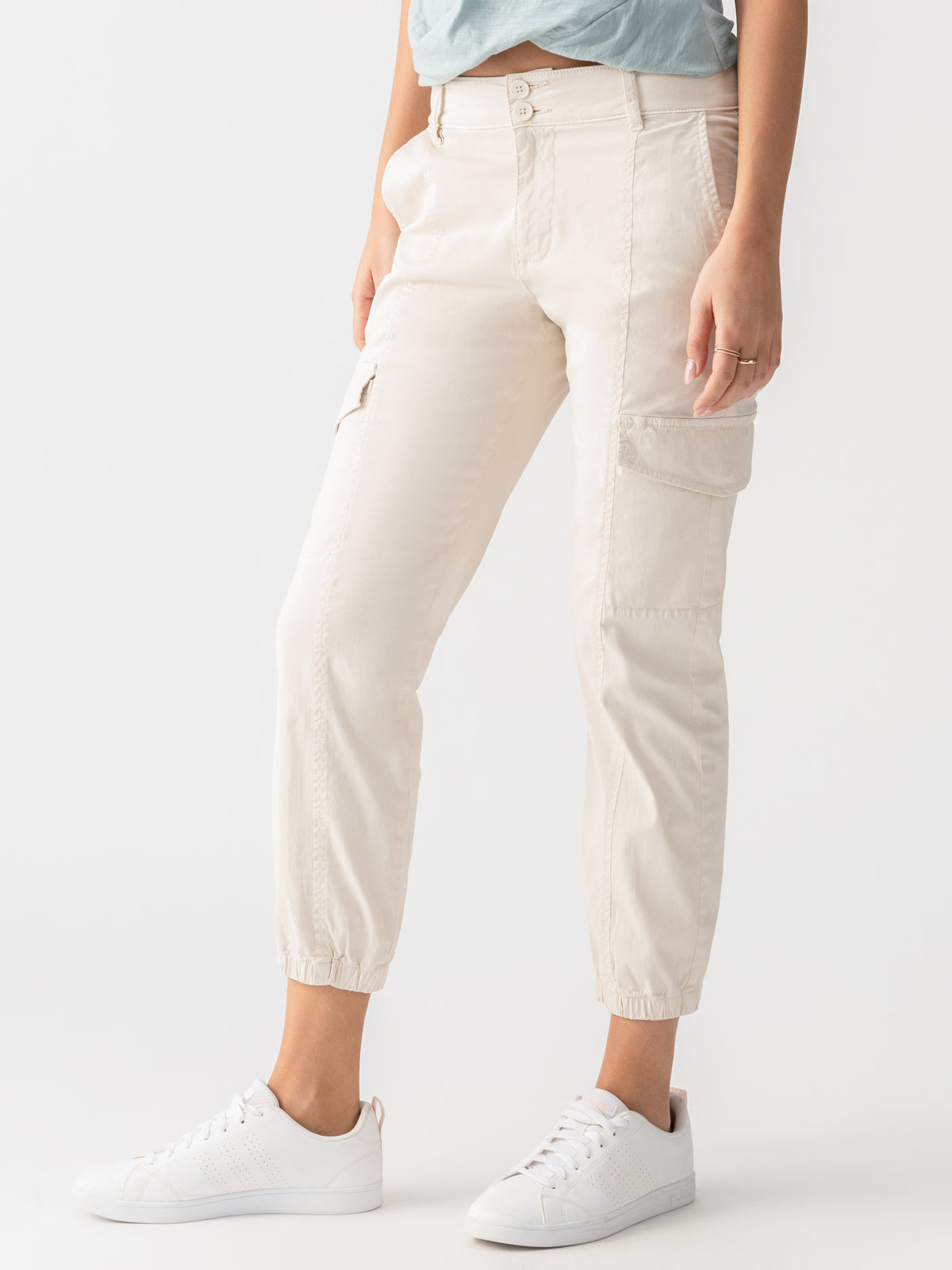 A person is wearing the Rebel Pant Crystal Stone from Sanctuary Clothing, which are light beige cargo pants with multiple pockets and cinched ankles. The person is also sporting white sneakers with a subtle pattern. Only the lower half of the body is visible, set against a neutral background.