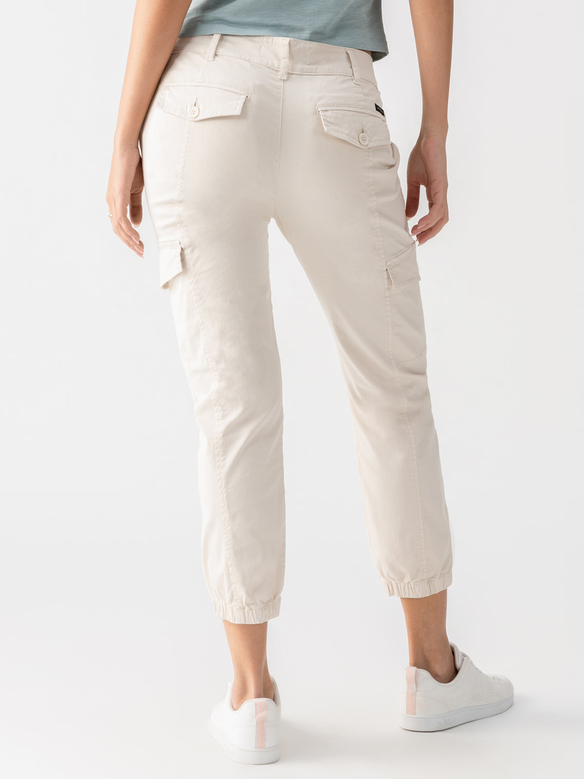 A person is shown from the back, wearing Sanctuary Clothing's Rebel Pant Crystal Stone, which are light beige cargo pants cropped above the ankles with elastic cuffs. The pants feature buttoned pockets on the back and sides. They are also wearing white sneakers with pink accents and a greenish-gray top.