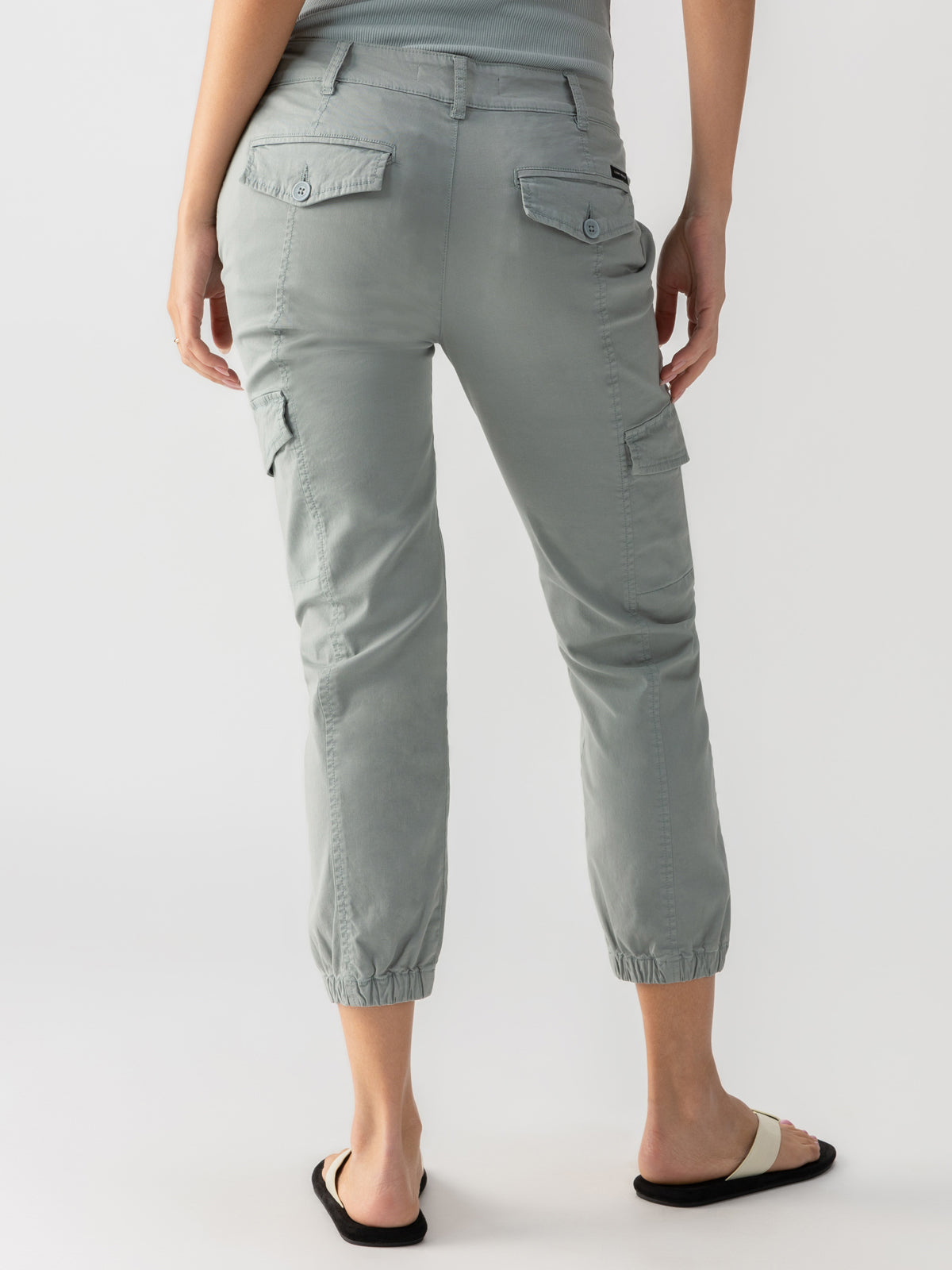 A person is seen from the back, sporting the Rebel Pant Eucalyptus by Sanctuary Clothing, which features light gray cargo pants with buttoned pockets and elastic cuffs at the ankles. They are also wearing a matching light gray top and black and white slide sandals.