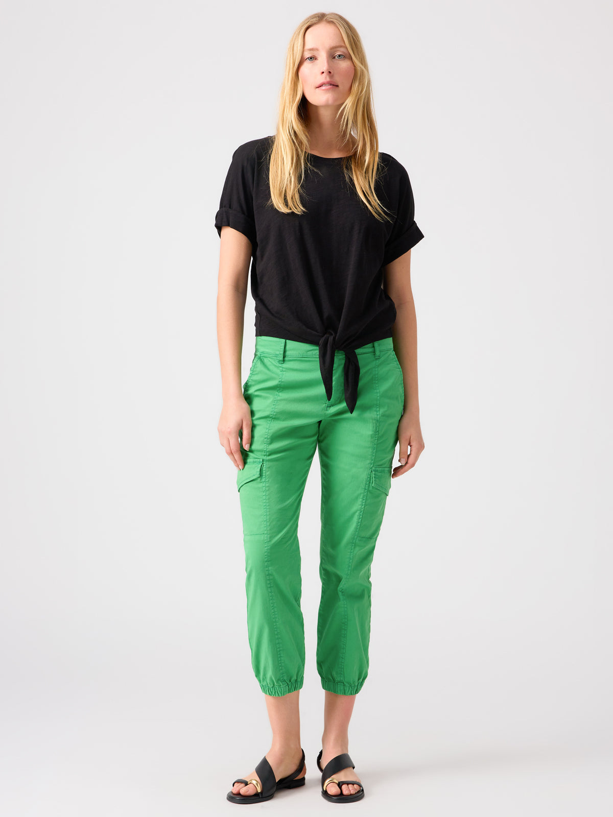 A person with long blonde hair wearing a black short-sleeve shirt tied at the front, Sanctuary Clothing's Rebel Pant Green Goddess with elastic cuffs, and black sandals stands against a plain white background.