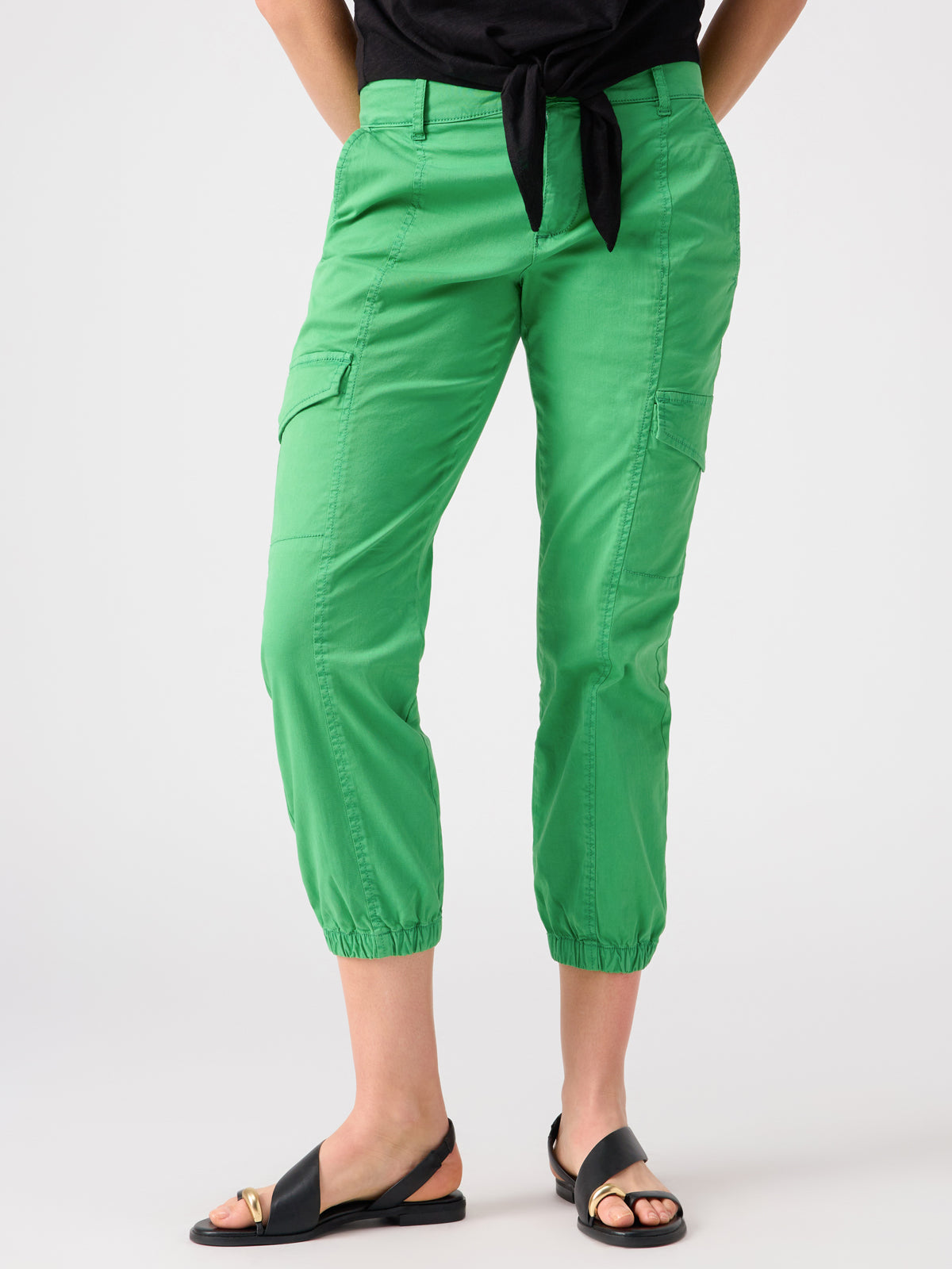 A person wearing Sanctuary Clothing's Rebel Pant Green Goddess with gathered cuffs, a black top featuring a waist tie, and black open-toe sandals adorned with gold accents stands against a plain white background, hands casually resting in their pockets.