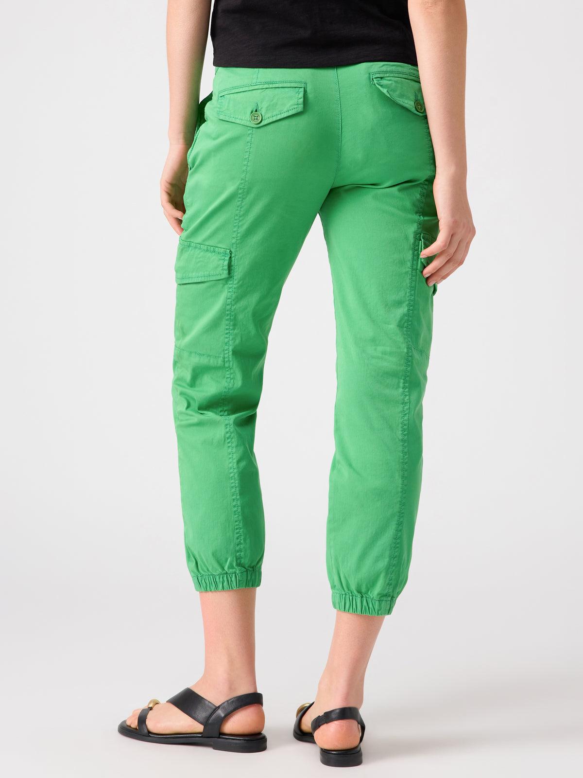 A person wearing the Rebel Pant Green Goddess cargo pants by Sanctuary Clothing with multiple pockets and elastic cuffs, paired with black sandals, stands against a plain background, facing away from the camera. Their upper body is partially visible, clothed in a black top.