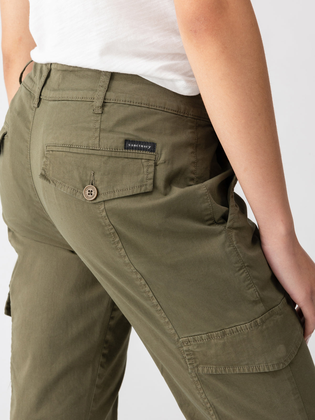 Close-up of a person wearing the Rebel Pant Hiker Green from Sanctuary Clothing, featuring multiple pockets, including a buttoned back pocket. They are also wearing a white t-shirt. The neutral background emphasizes the details and stitching of the pants.