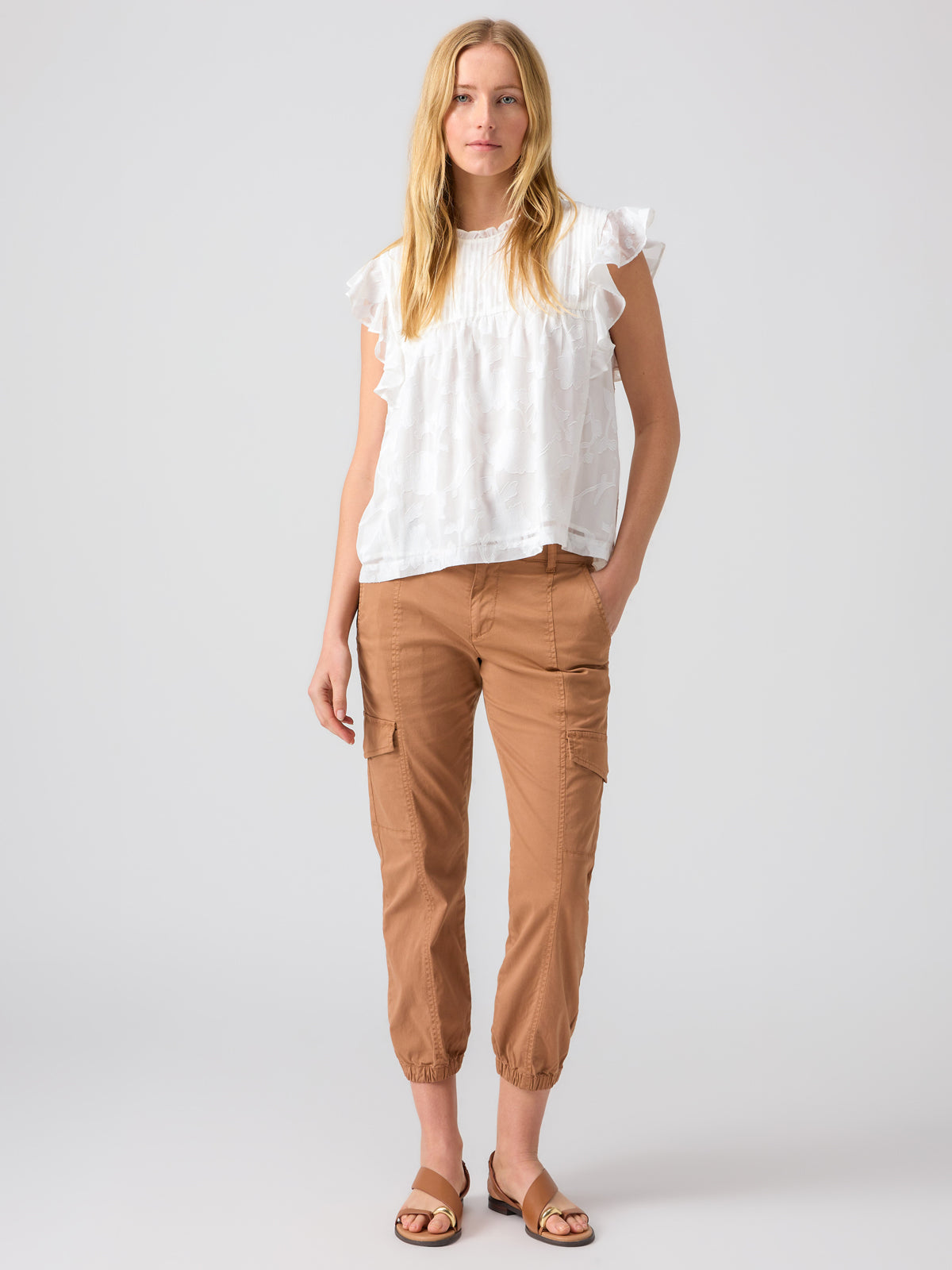 A woman with long blonde hair stands against a plain background. She is wearing a white blouse with ruffled sleeves, Sanctuary Clothing's Rebel Pant in Mocha Mousse, and tan sandals. Her expression is neutral, and her arms are relaxed at her sides.