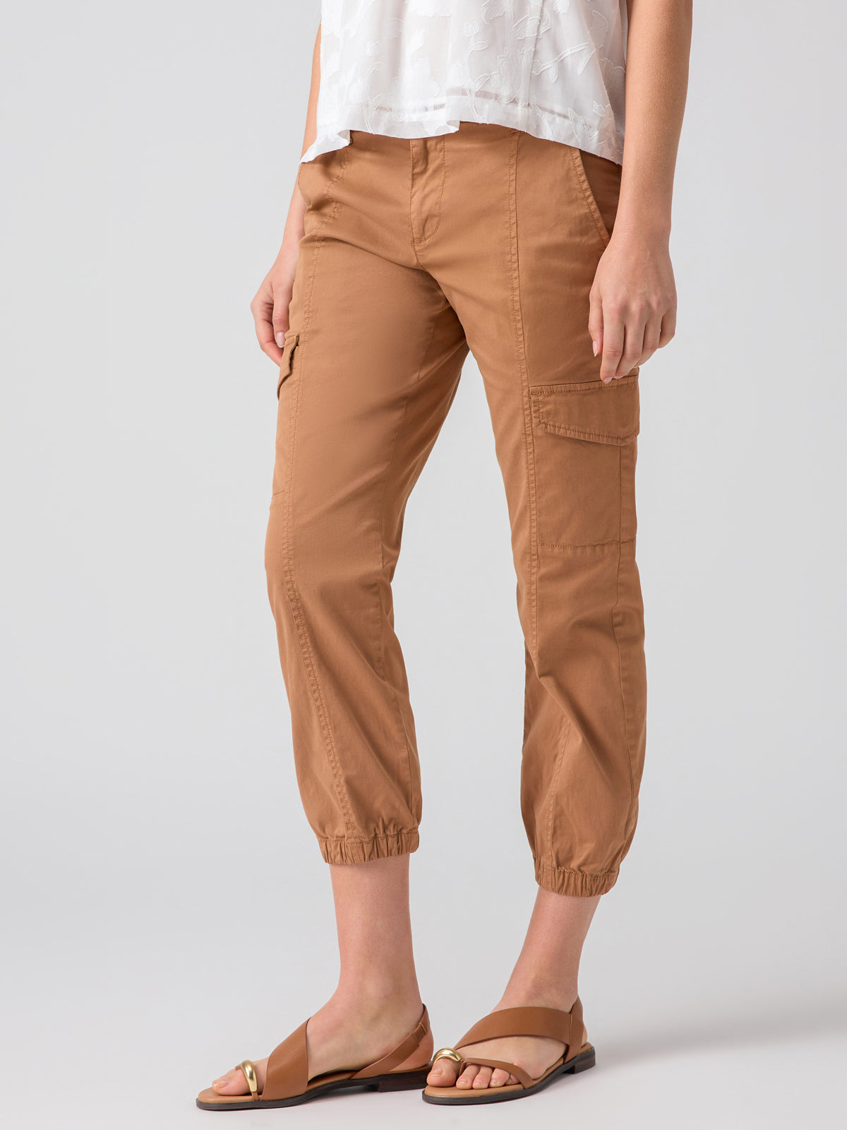 A person is wearing the Rebel Pant Mocha Mousse by Sanctuary Clothing along with a white patterned top. They are also wearing brown open-toe sandals with gold accents. The background is plain gray, and the photo captures the person from the shoulders down.