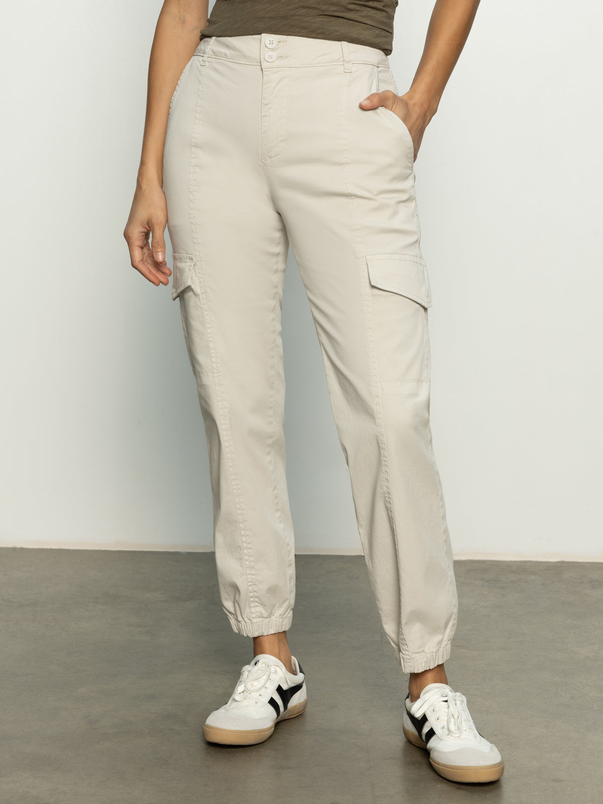 A person in Sanctuary Clothing's rebel pant pumice stone cargo pants, featuring elastic cuffs, a dark top, and white sneakers stands against a plain background with their left hand in the pants pocket.