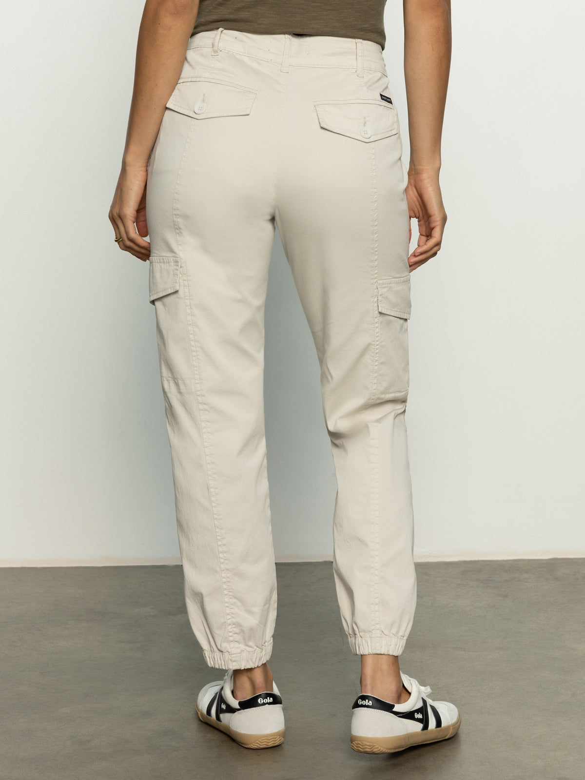 A person wearing Sanctuary Clothing's rebel pant pumice stone stands on a gray floor. The light cargo pants have flap pockets on the back and sides with elastic cuffs, paired with striped sneakers and a dark green top.