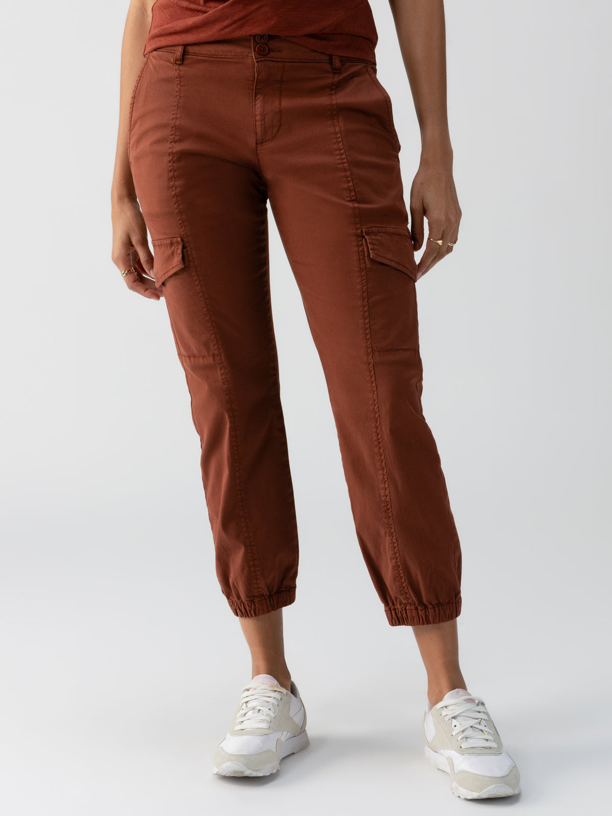 A person stands against a plain background wearing Sanctuary Clothing's Rebel Pant in Rich Clay, featuring thigh pockets and elastic cuffs. They are also dressed in a matching rust-colored top and white sneakers. The image shows only the person from the shoulders down.