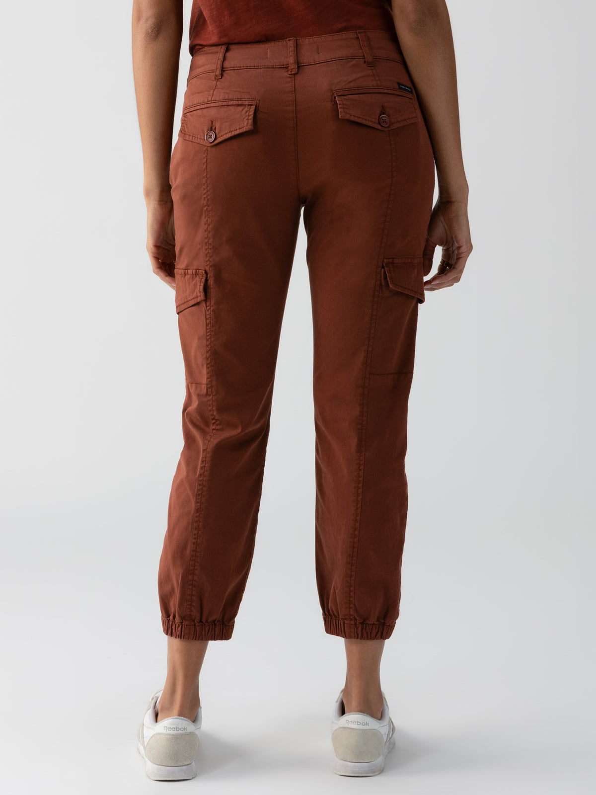 Rear view of a person dressed in the Rebel Pant Rich Clay from Sanctuary Clothing. These rust-colored cargo pants feature multiple pockets, including button-flap pockets on the back and side cargo pockets. The person is also wearing a matching rust-colored top and white sneakers. The hems of these stylish pants are elasticated, set against a plain, light-colored background.