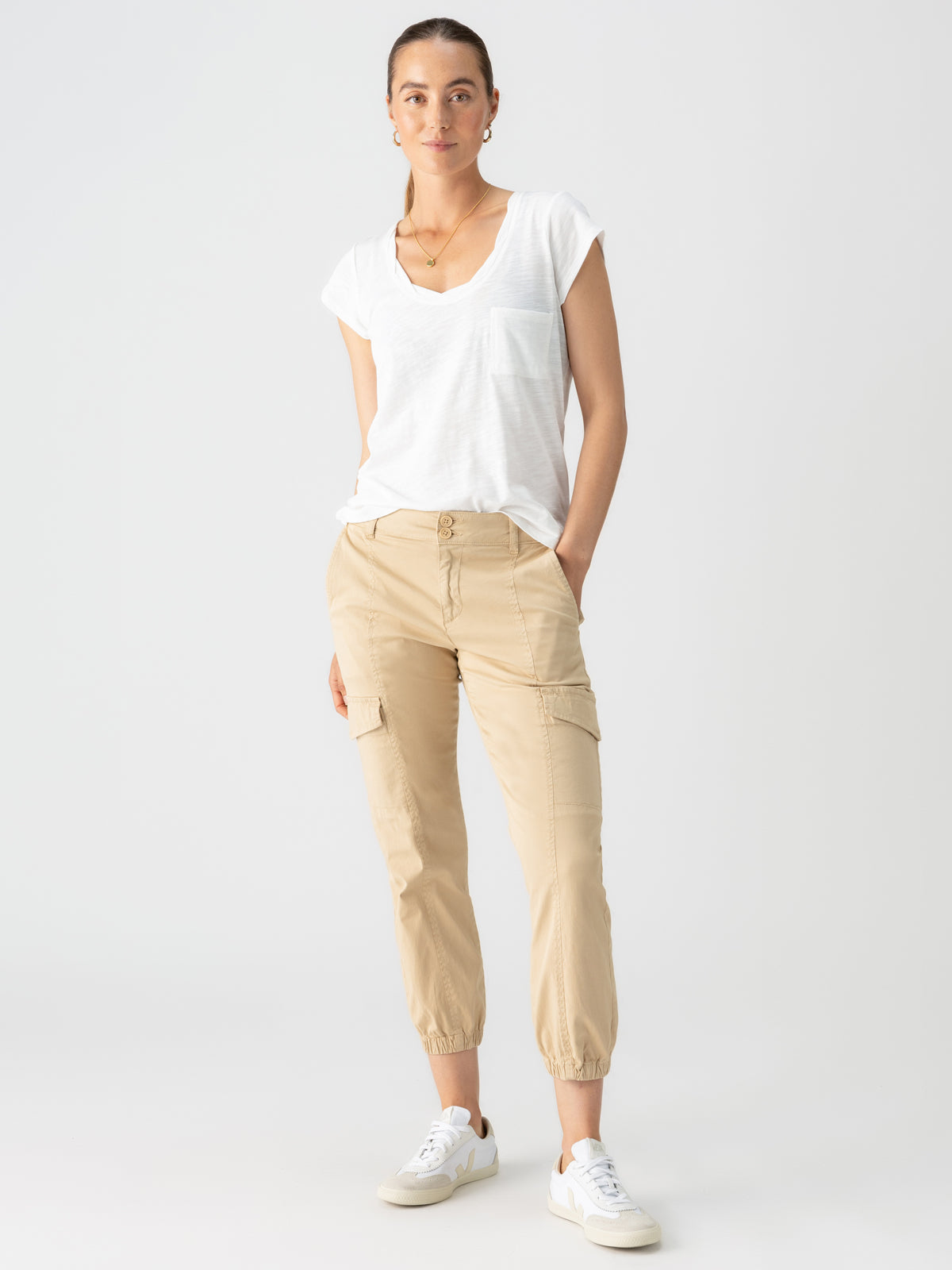 A person stands confidently against a plain background, wearing a casual outfit consisting of a white short-sleeved t-shirt with a pocket, the Rebel Pant True Khaki by Sanctuary Clothing, and white sneakers. They have their hair tied back and are accessorized with a necklace and small earrings.