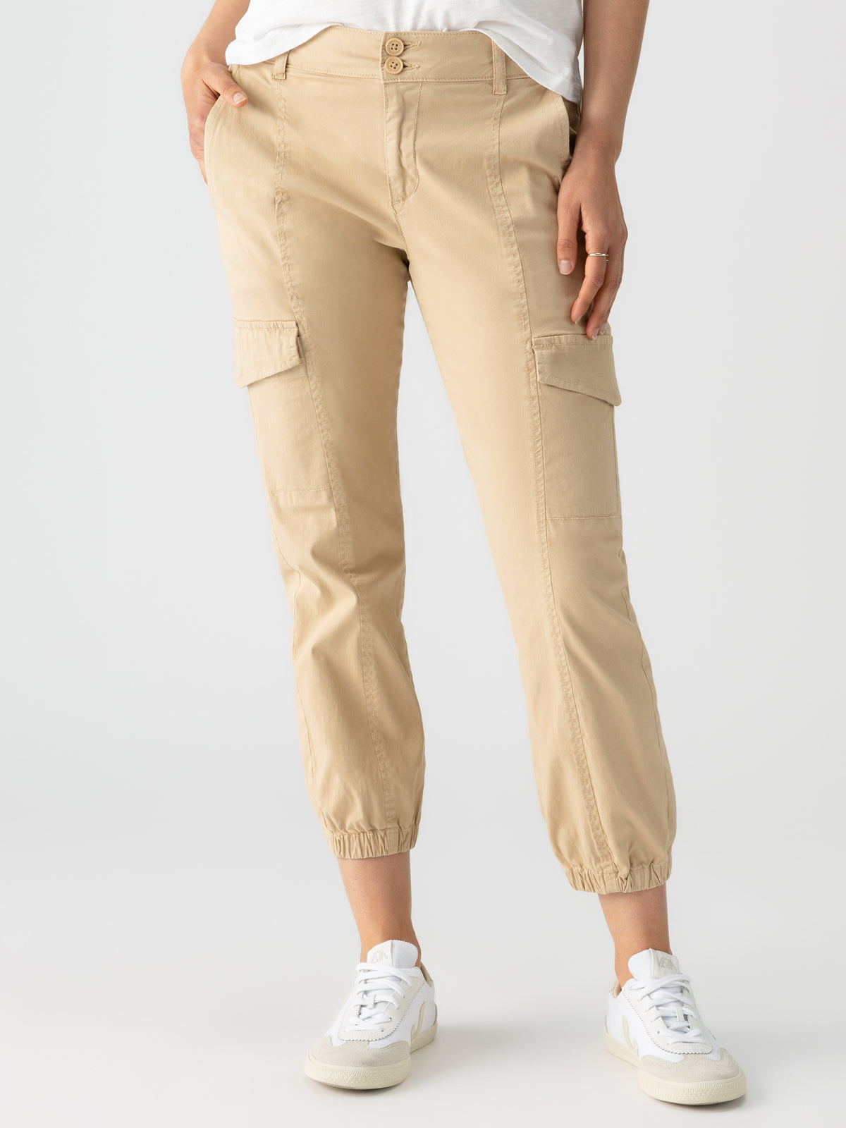 A person wearing Sanctuary Clothing's Rebel Pant in True Khaki stands against a plain white background. The pants, featuring side pockets and cuffed ankles, are paired with white sneakers. One hand is in a pocket while the person wears a partially visible white top.