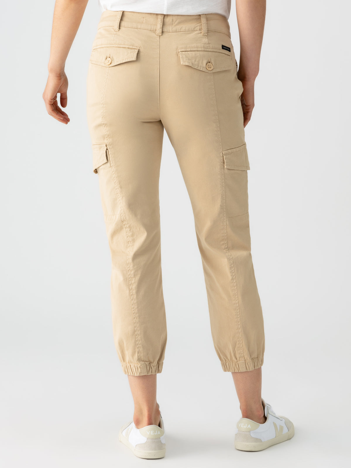 A person wearing Sanctuary Clothing's Rebel Pant True Khaki, featuring elastic cuffs and side and back pockets with button flaps, along with white sneakers. The individual is standing with their back facing the camera against a light, plain background.