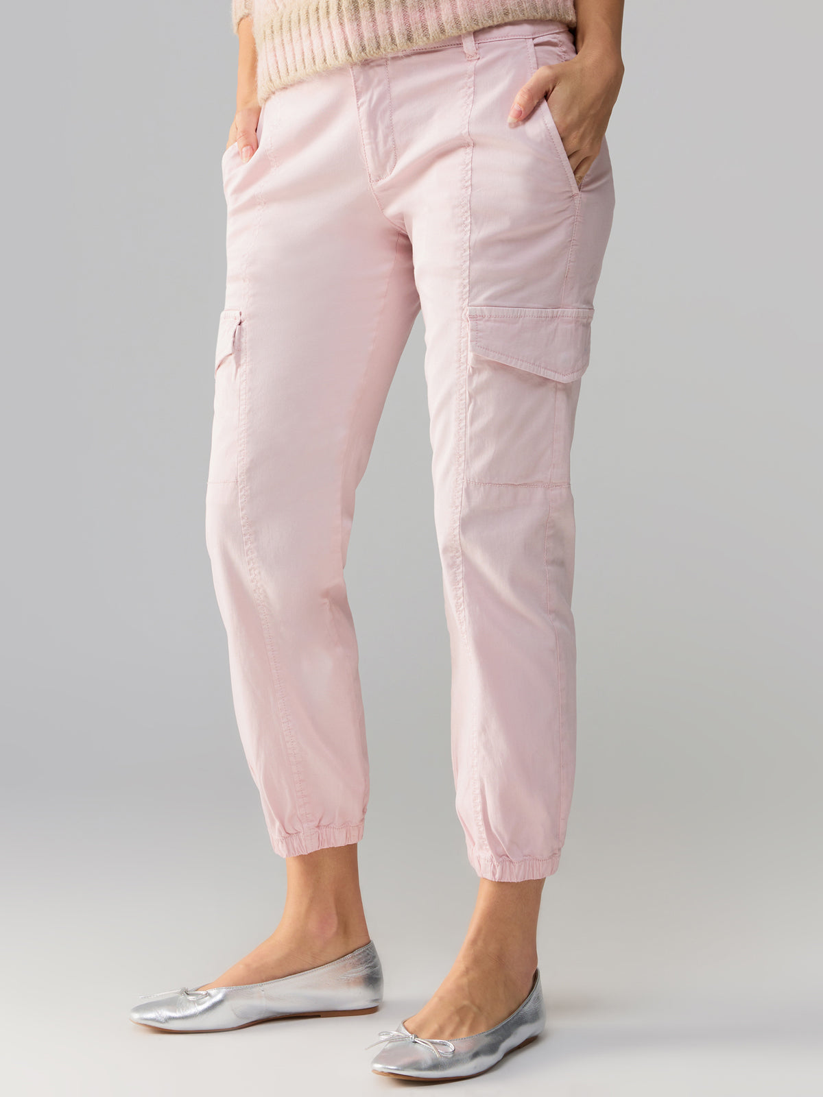 A person is dressed in Sanctuary Clothing's Rebel Pant Washed Pink No 3, featuring side pockets and gathered ankles. They are also wearing a light-colored knit sweater paired with metallic silver ballet flats. With their hands tucked into the pockets, they pose against a neutral background.