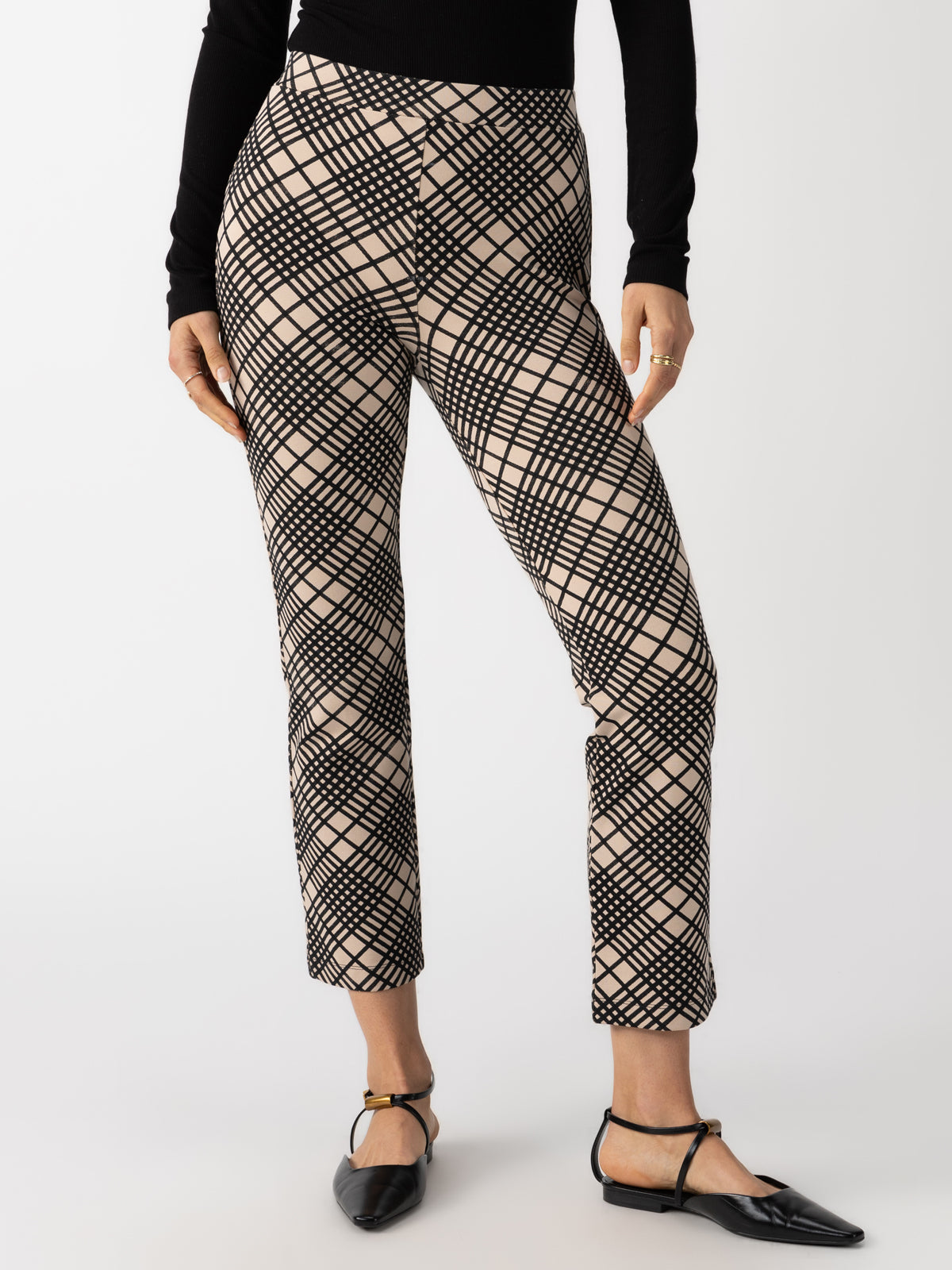 A person wearing the Carnaby Kick Crop Legging Graphic Plaid pants by Sanctuary Clothing in a black and beige plaid pattern, paired with a black long-sleeve top. The person's hands are by their sides, and they are wearing black flats with ankle straps. The background is plain white.