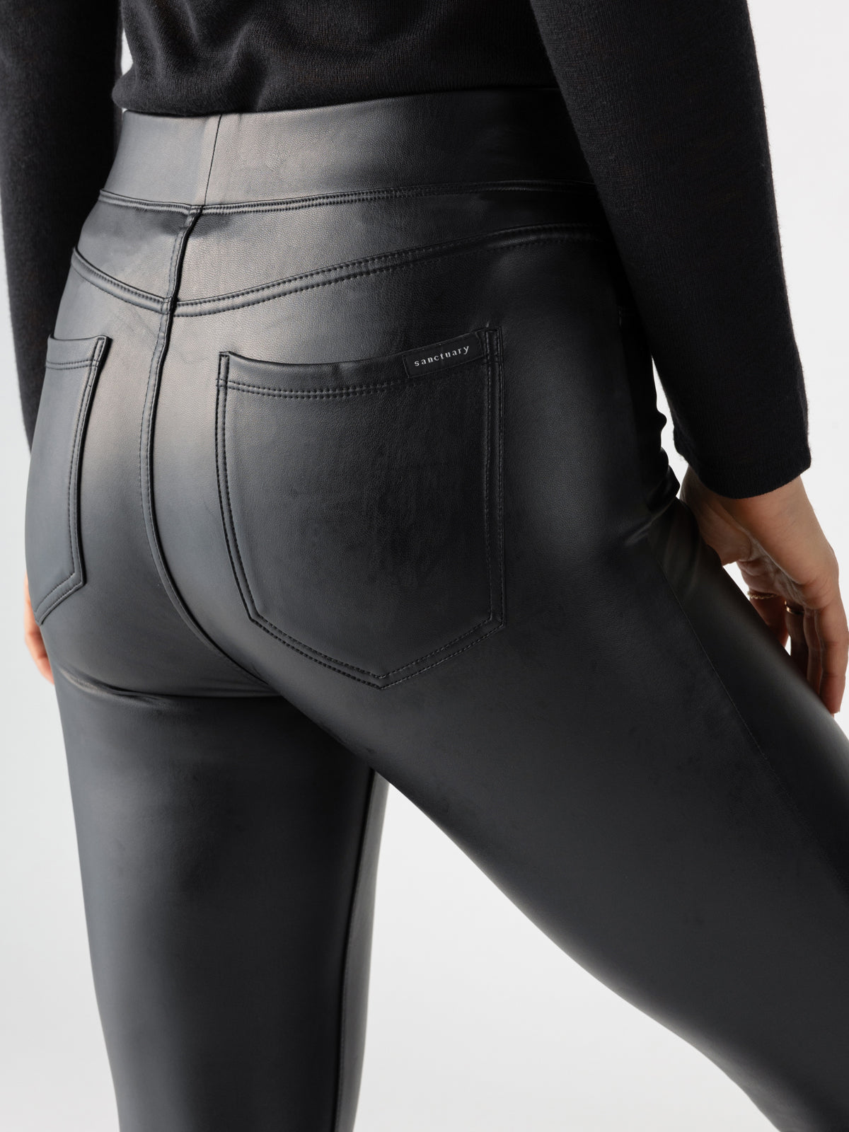 A person dressed in Runway Legging Slick Black from Sanctuary Clothing and a black long-sleeve top is captured from the waist down. The focus is on the pants' back, highlighting the pockets and the glossy texture.