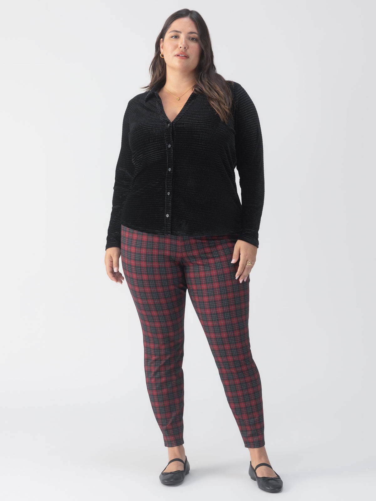 In front of a simple white backdrop, an individual is dressed in a black long-sleeve button-up shirt and Sanctuary Clothing's Runway Legging Mars Red Check from the Inclusive Collection. They complete the look with black flats.