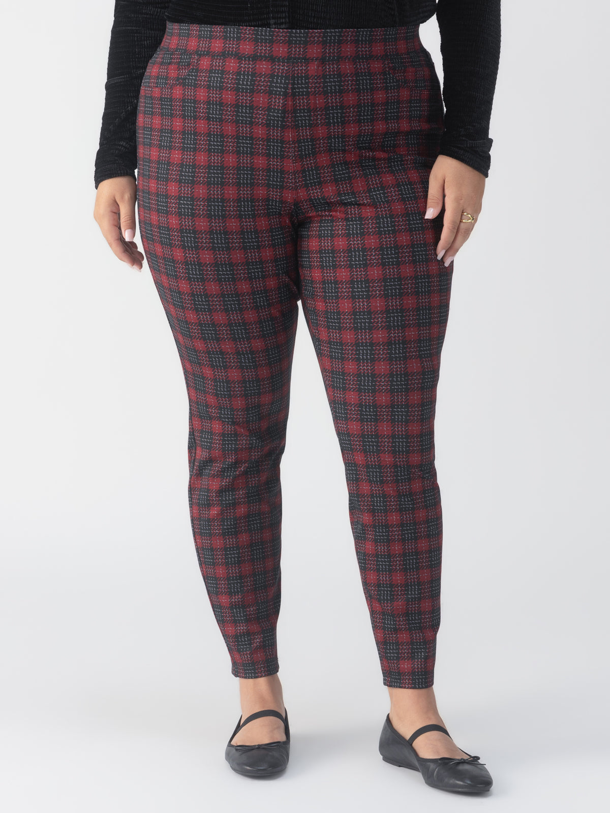 A person wearing the Sanctuary Clothing Runway Legging Mars Red Check from the Inclusive Collection stands against a plain background. They are also wearing a black textured long-sleeve top and black flats, with their arms resting at their sides.