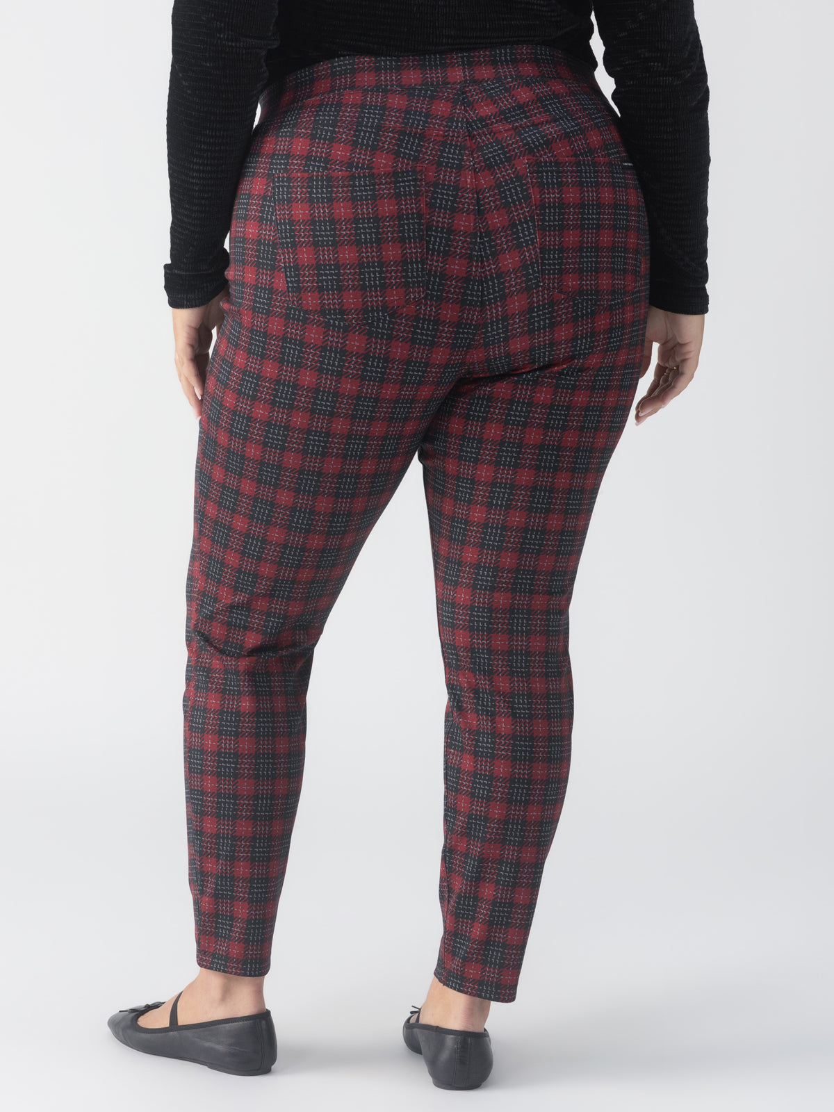 A rear view of a person wearing Sanctuary Clothing's Runway Legging in Mars Red Check from the Inclusive Collection. They are paired with a black long-sleeve top and dark loafers, standing against a light background with hands by their sides.