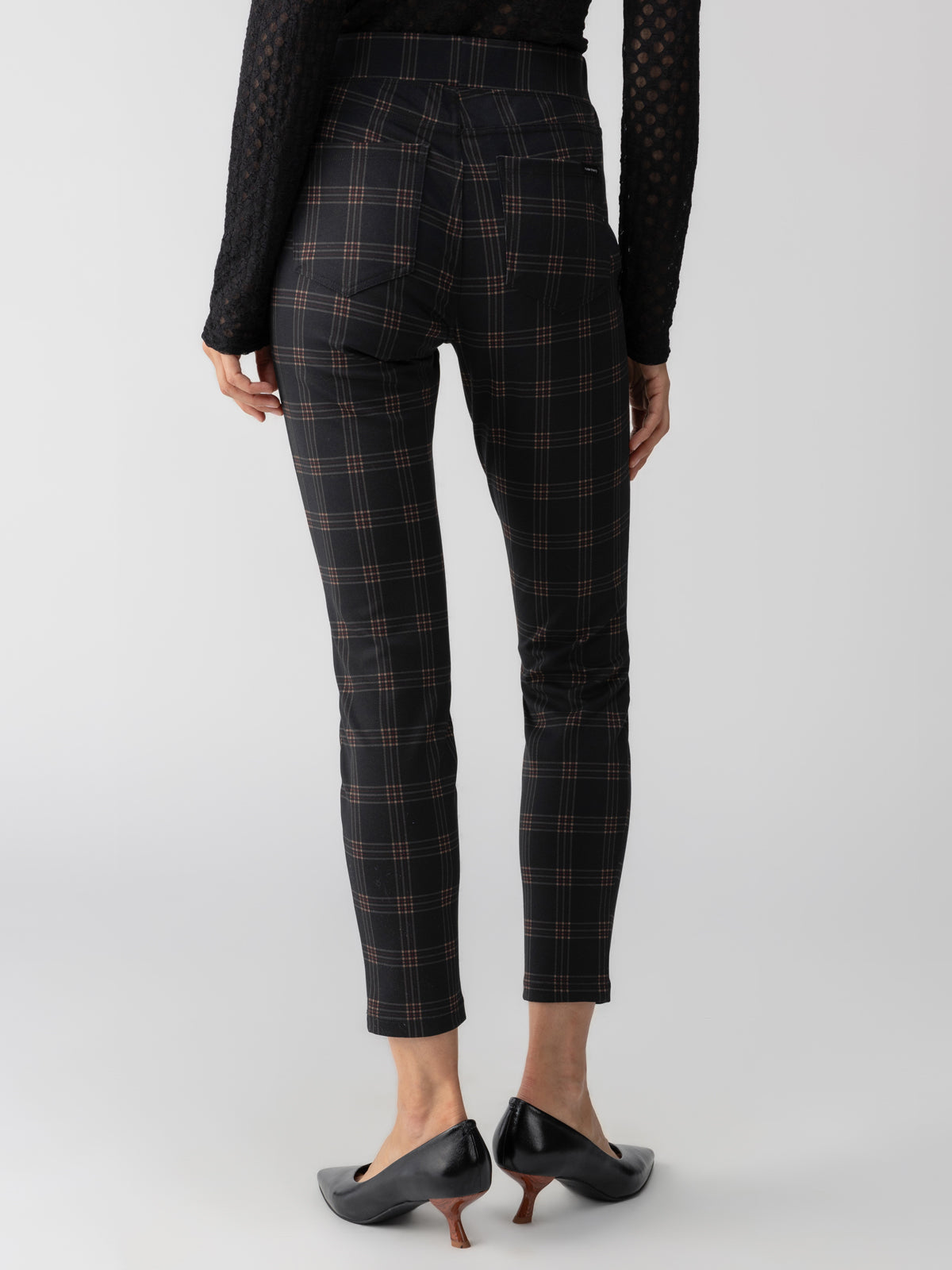 The image features the back view of a person dressed in Sanctuary Clothing's Runway Legging Garnet Plaid high-waisted trousers, alongside a black long-sleeved top and black closed-toe heeled shoes. The person's hands are resting by their sides against a plain white background.